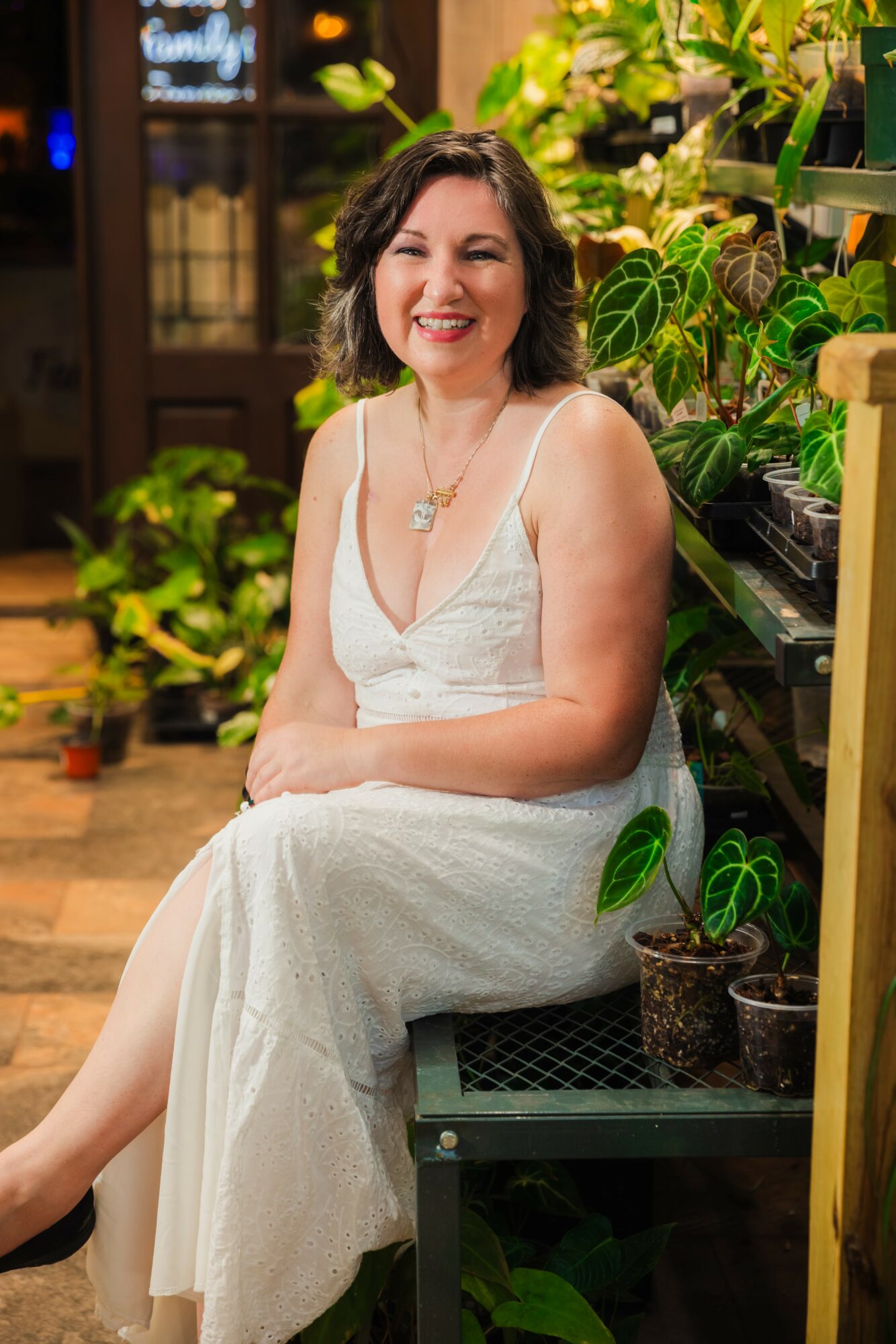 Woman in white dress sitting among potted plants, smiling, indoors with wooden furniture and greenery.