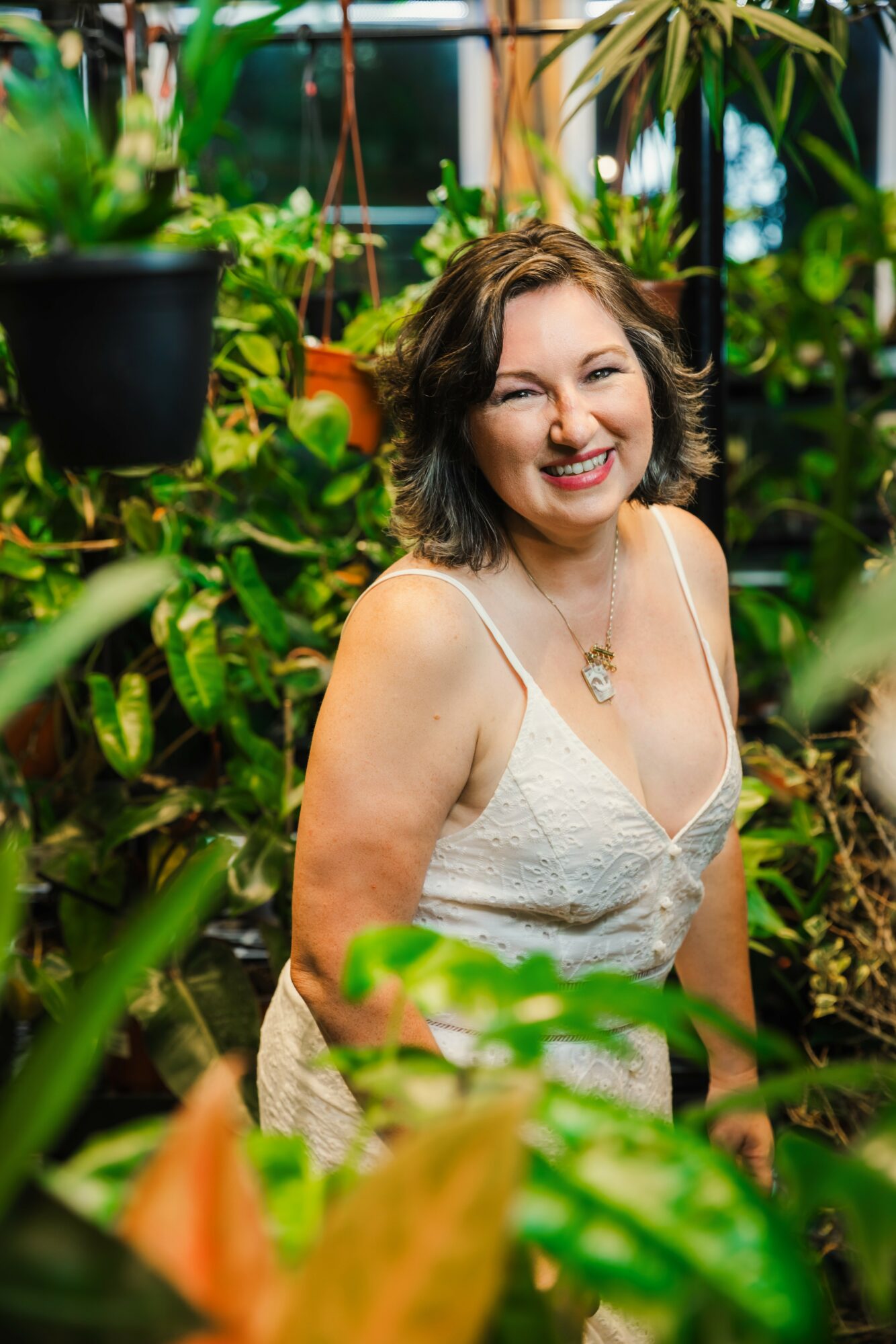 Smiling woman with shoulder-length curly hair in a white dress surrounded by green plants in a greenhouse.