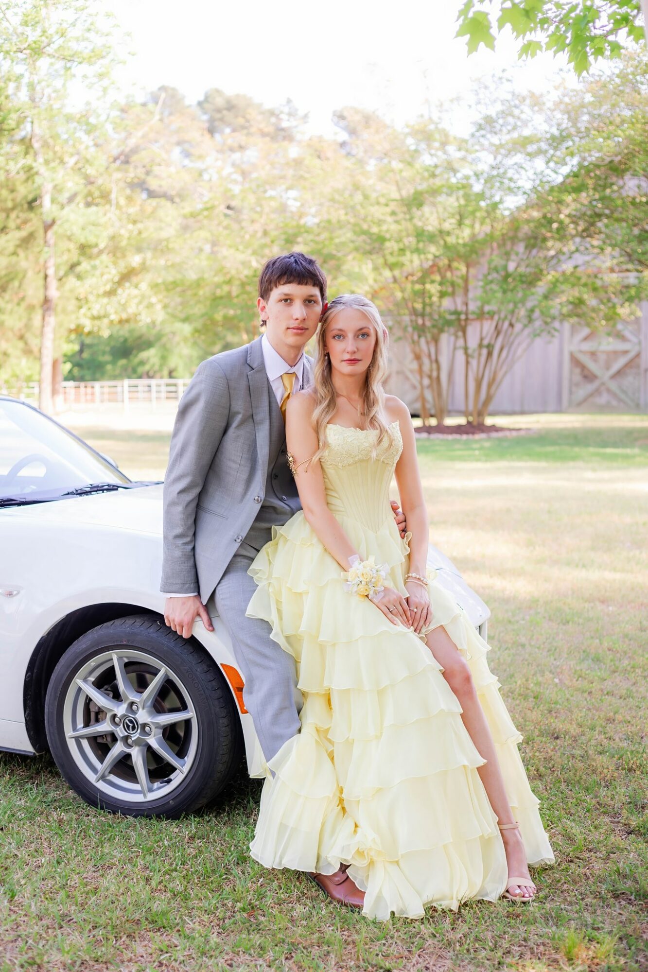 A young man in a gray suit and a young woman in a yellow dress stand outdoors near a white car, with trees in the background.