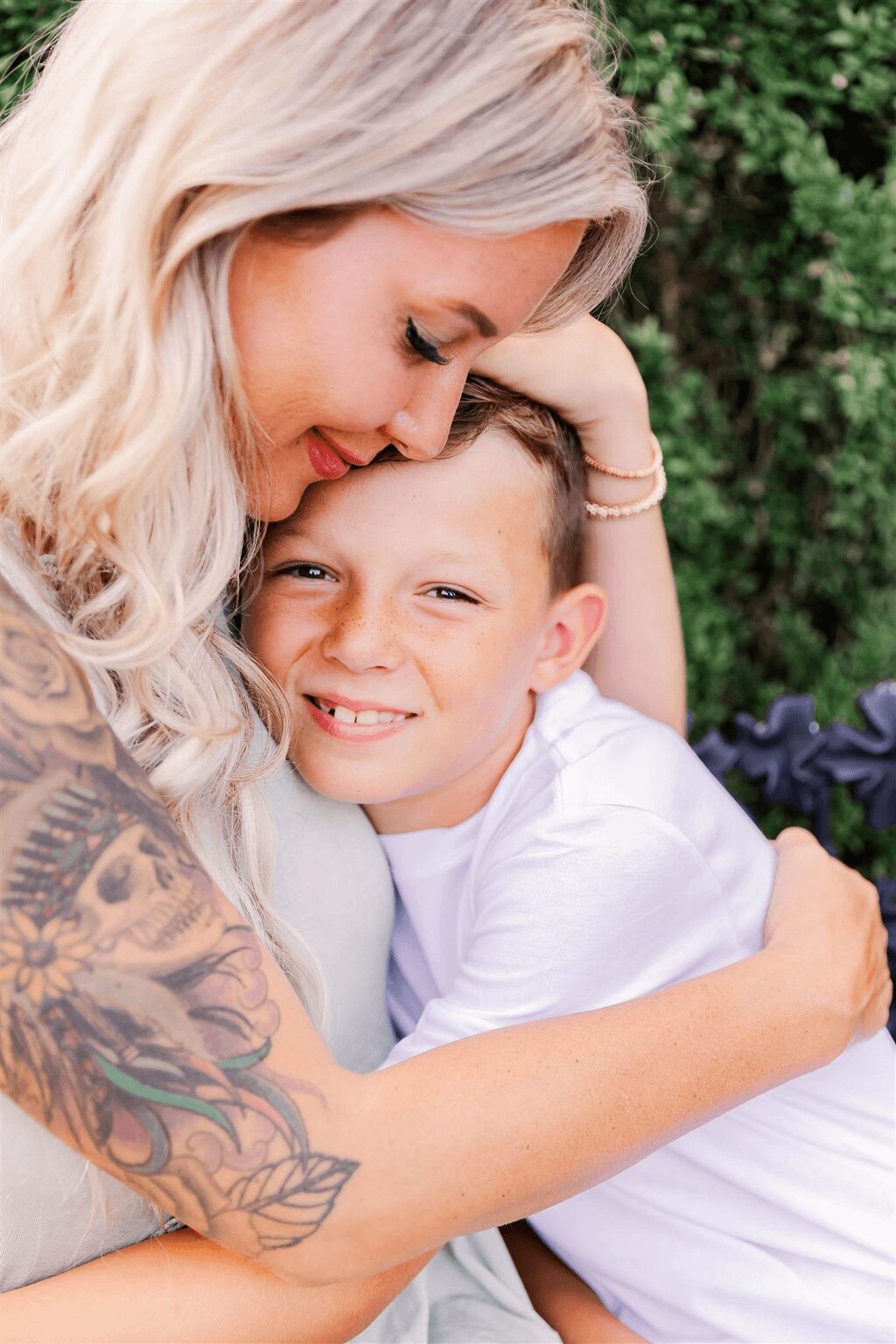 Woman and boy hugging outdoors, woman smiling, boy with a big smile, greenery in background.