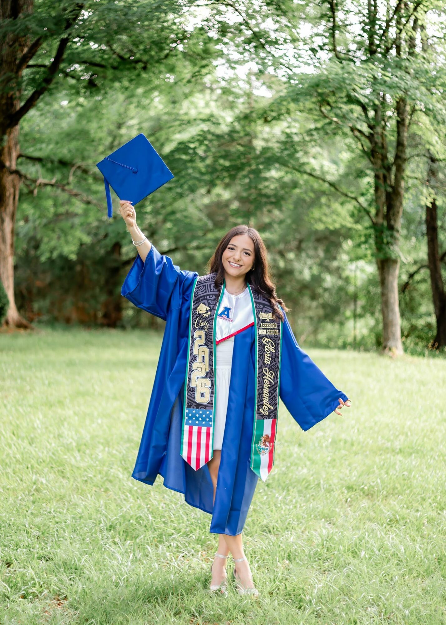 Young woman in graduation gown holding a diploma outdoors in a park with trees and grass.