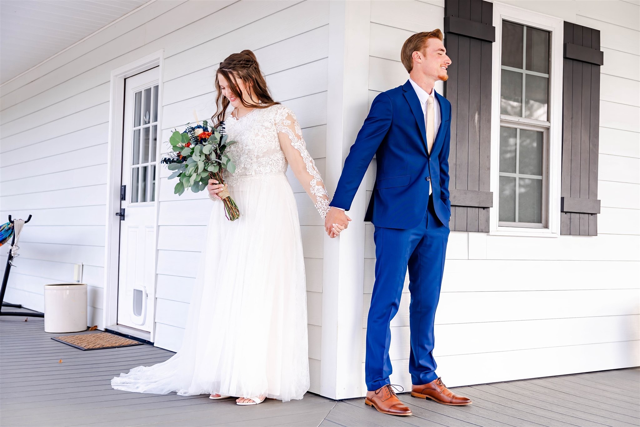 Bride and groom holding hands outside a white house, bride in a white dress holding a bouquet, groom in a blue suit.