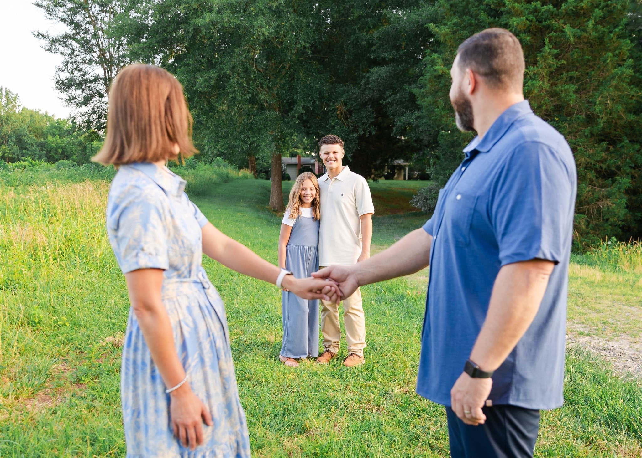 A woman and a man shake hands outdoors with two children in the background, trees and grass around them.