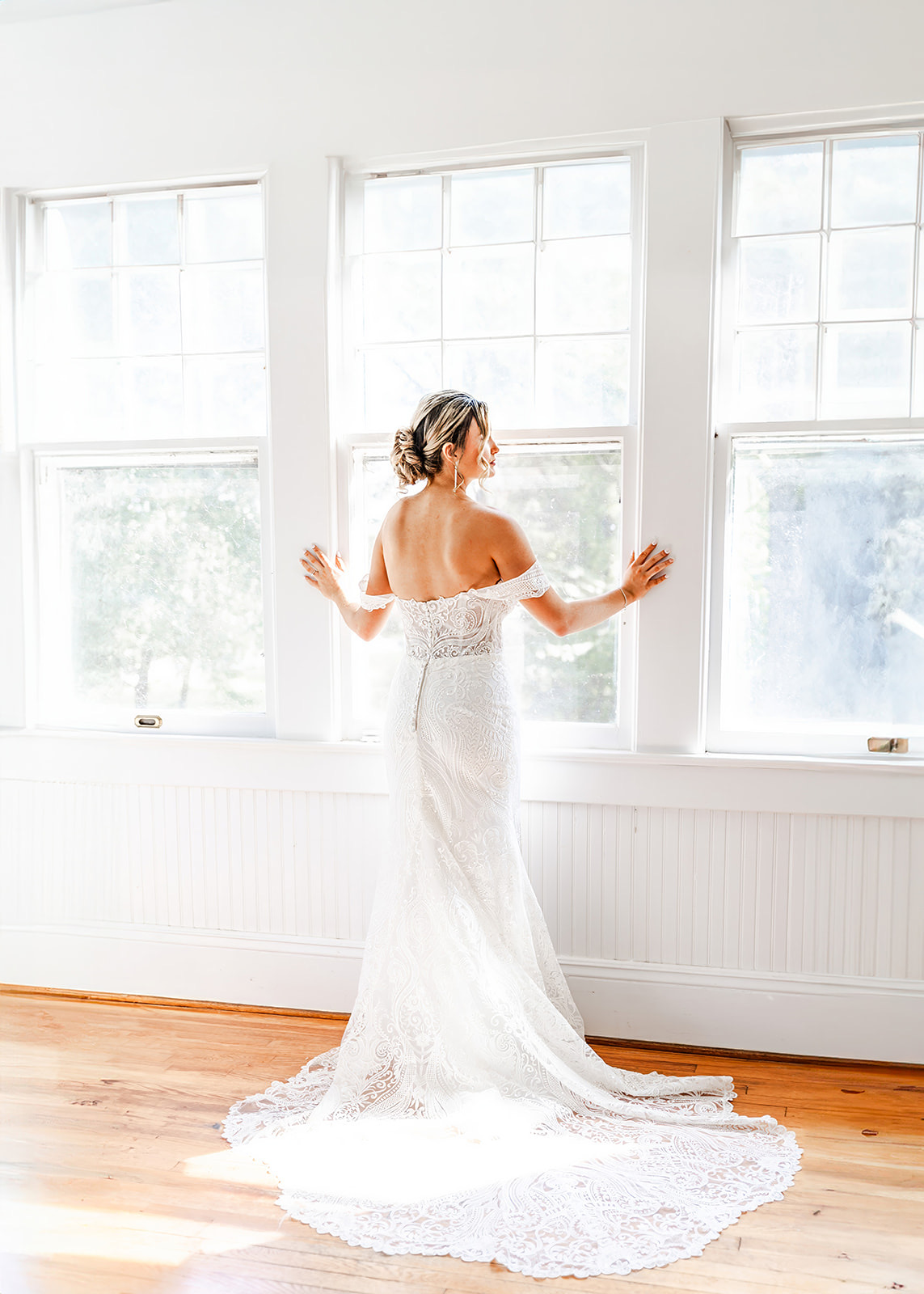 Woman in a white lace dress standing by three large windows, looking outside, with natural light filling the room.