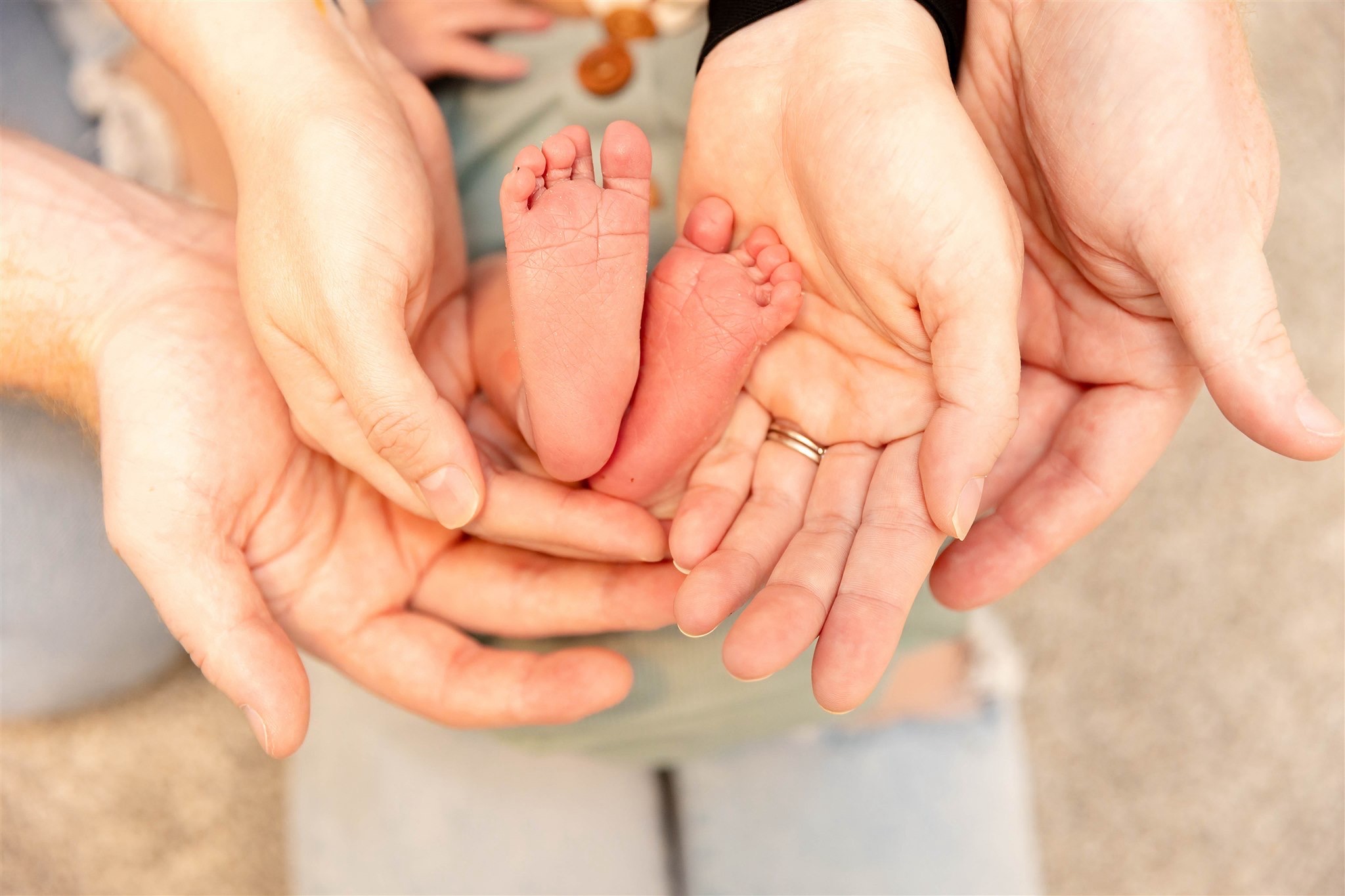 Multiple hands gently cradle tiny newborn feet and legs, forming a circle around the baby, on a neutral background.