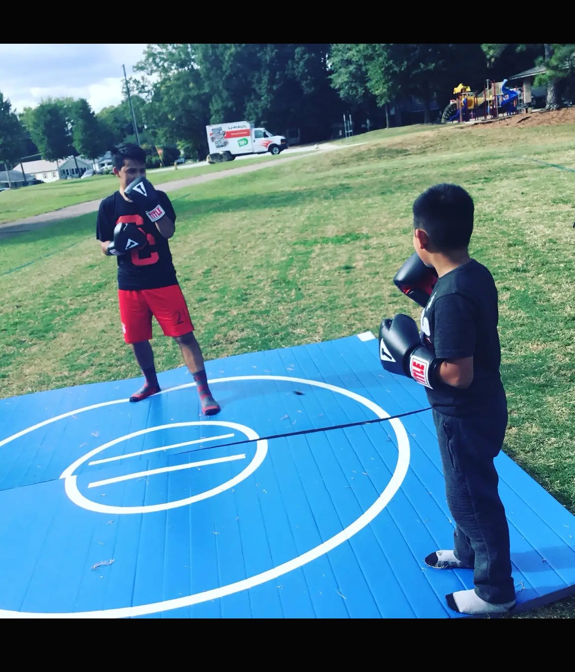 Two boys in boxing gloves stand on a blue outdoor boxing ring, facing each other, in a park with trees and a truck in the background.