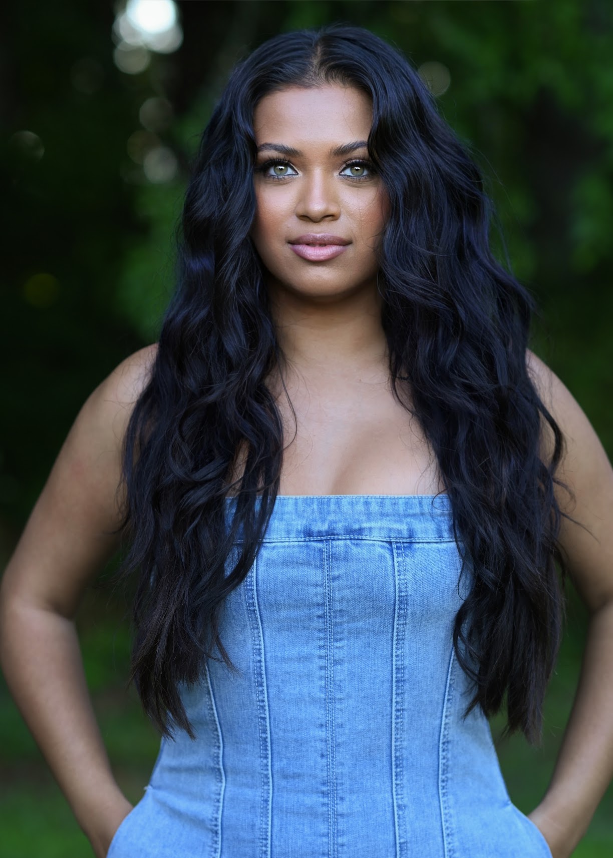Young woman with long wavy dark hair wearing a blue strapless top outdoors, green background.