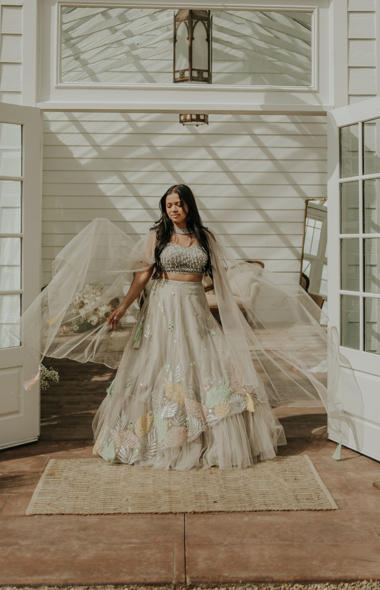 Woman in a long, flowing dress standing on a porch with open doors, sunlight casting shadows, and a lantern above.