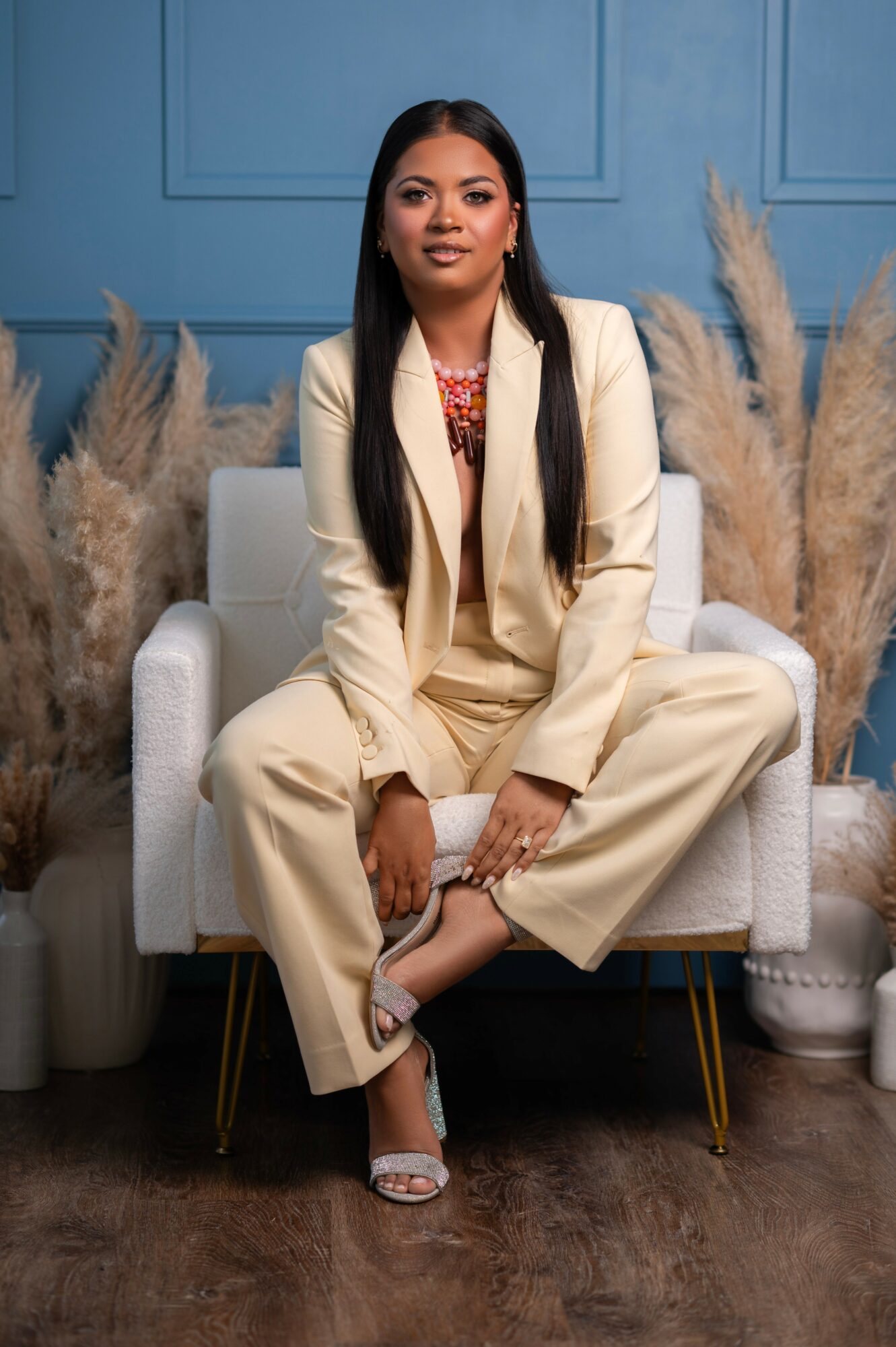 Woman sitting on a white armchair, wearing a cream-colored suit, with pampas grass in the background, blue wall, and wooden floor.