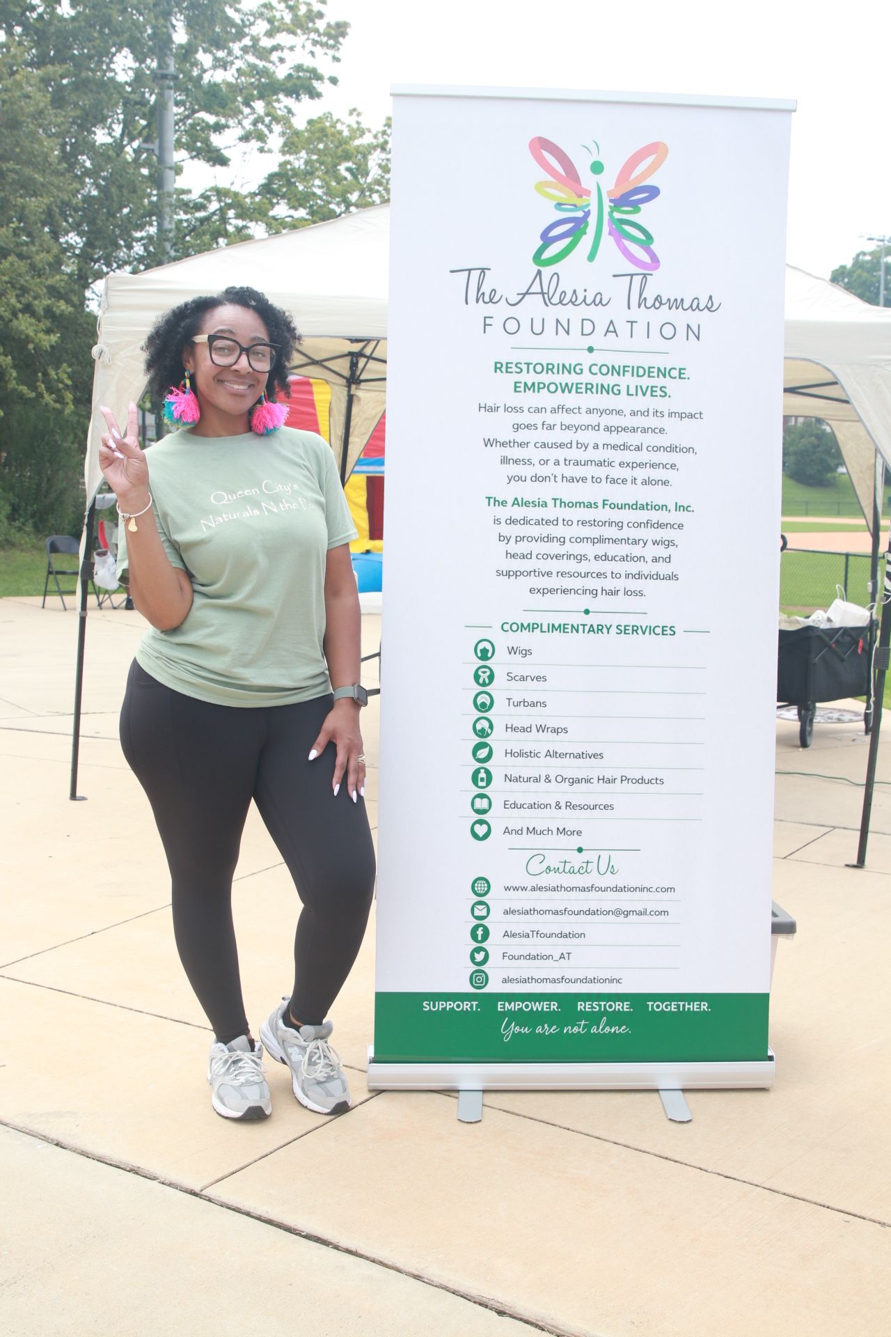Woman standing outdoors next to a large informational sign, smiling, with trees and a playground in the background.