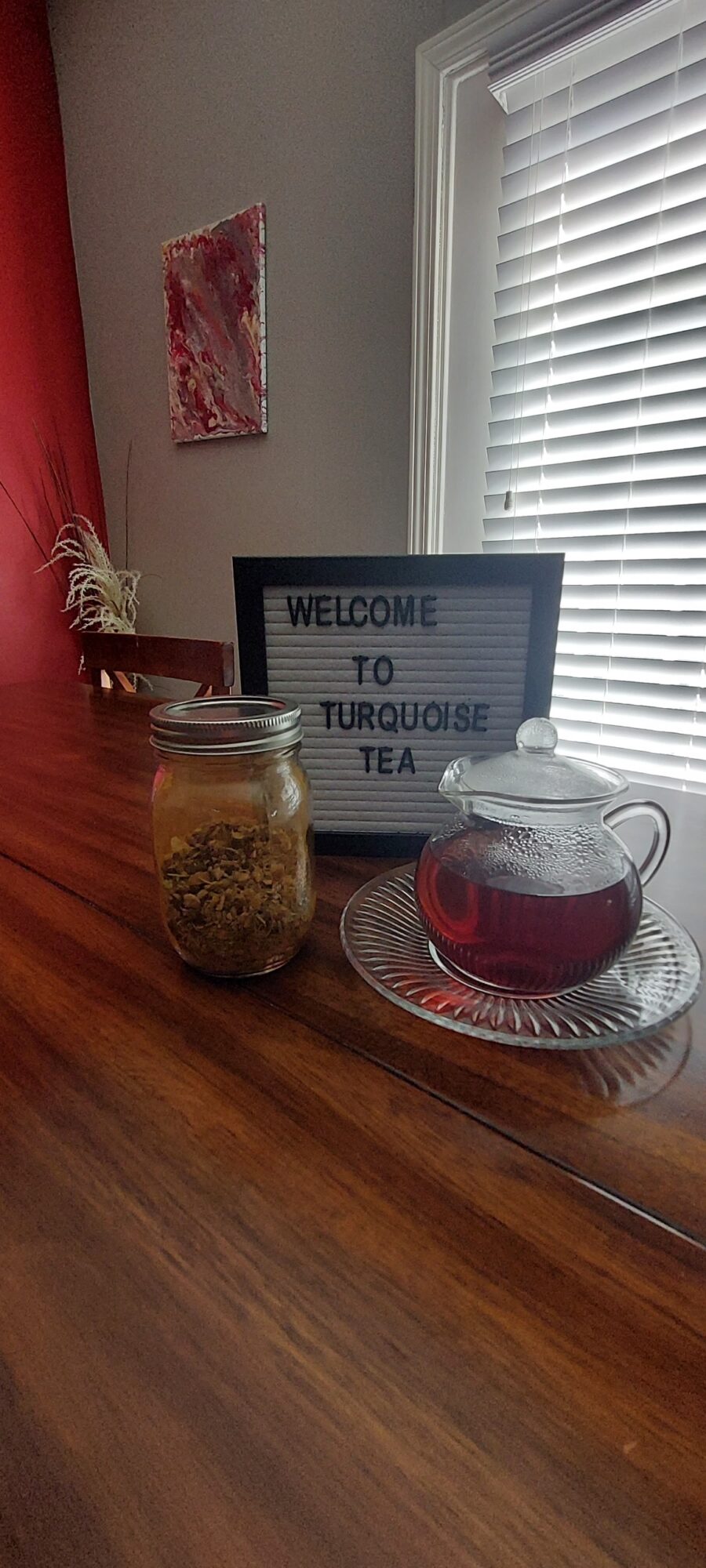 Tea setup with a glass teapot, a sign, and a jar on a wooden surface near a window.