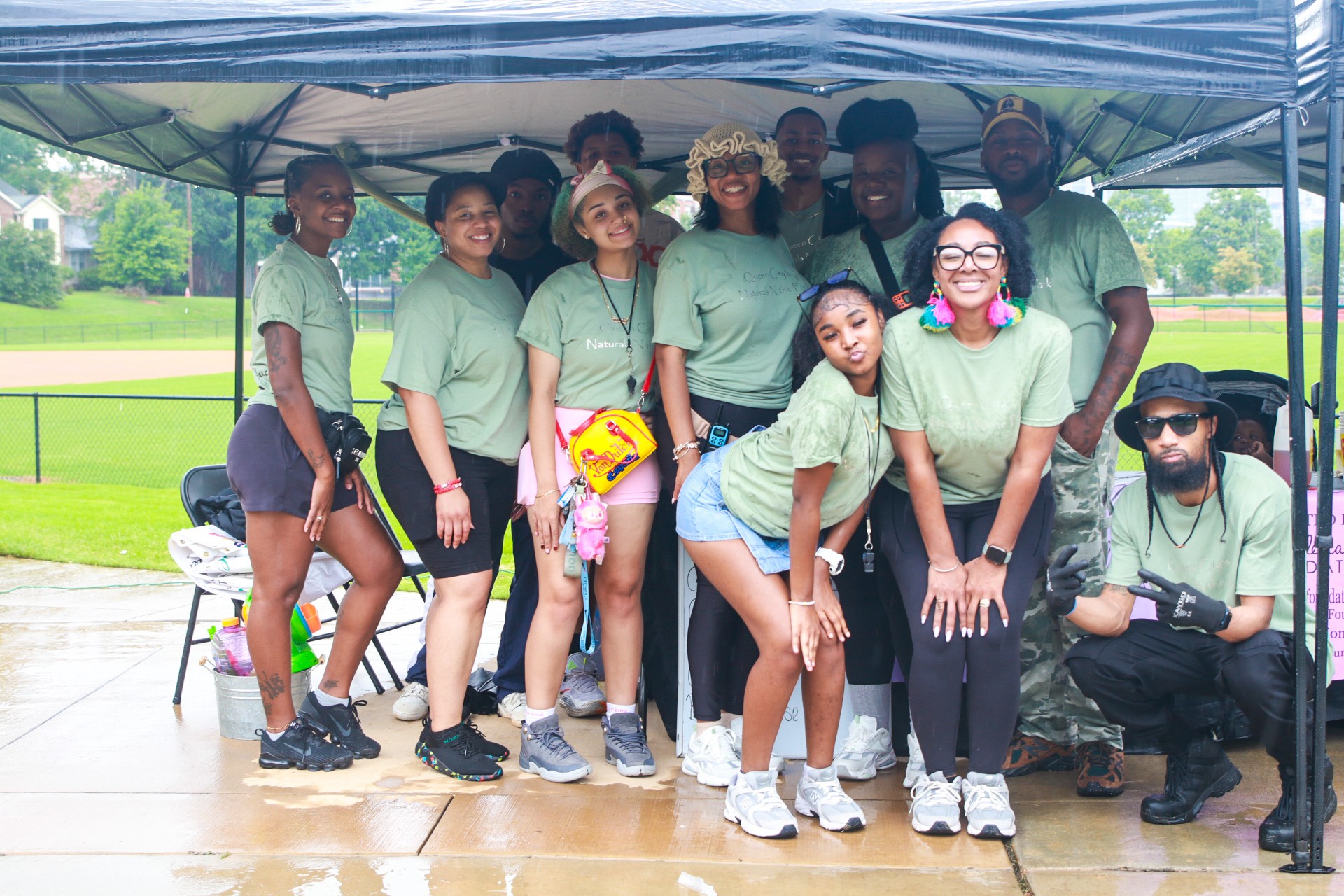 Group of people wearing matching green shirts under a canopy outdoors, some standing and some sitting, with a park background.