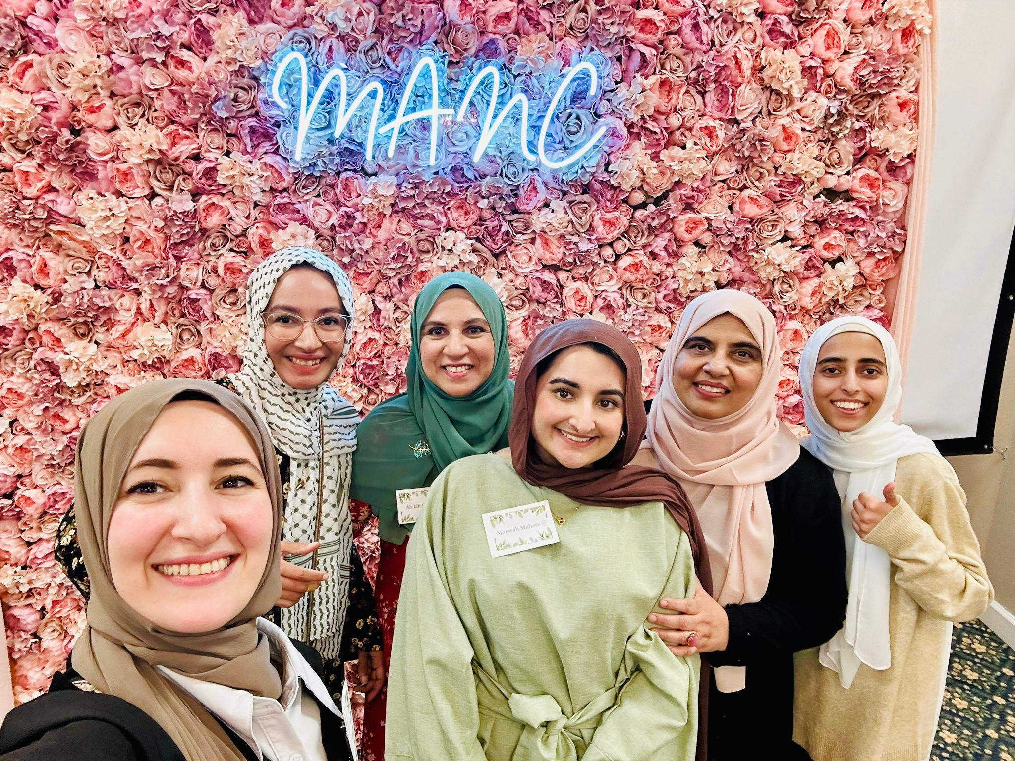 Six women smiling in front of a pink floral backdrop with a neon sign that says 'MANG'.