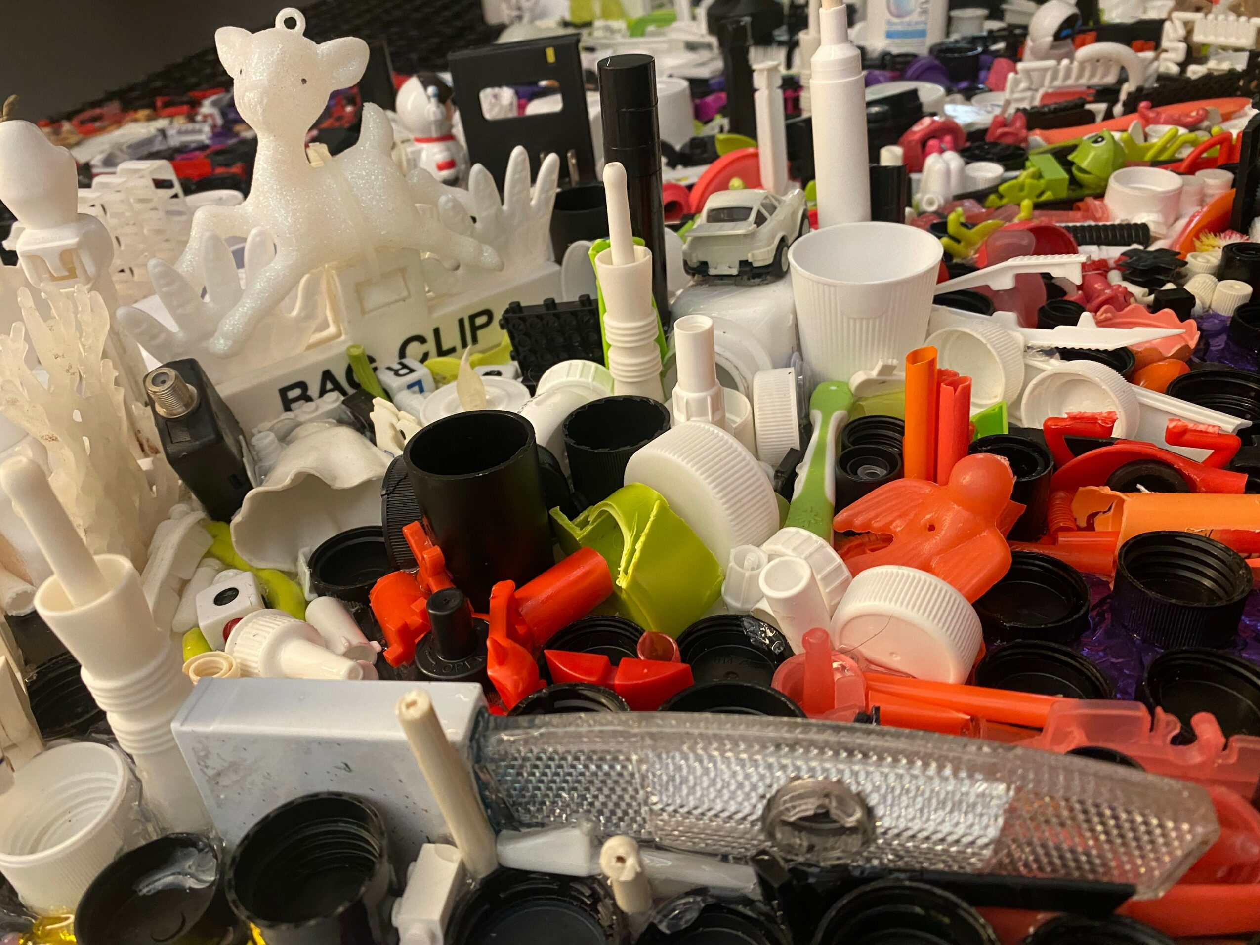 Pile of colorful plastic objects, bottles, and toys on a table, with a white toy giraffe in the background.