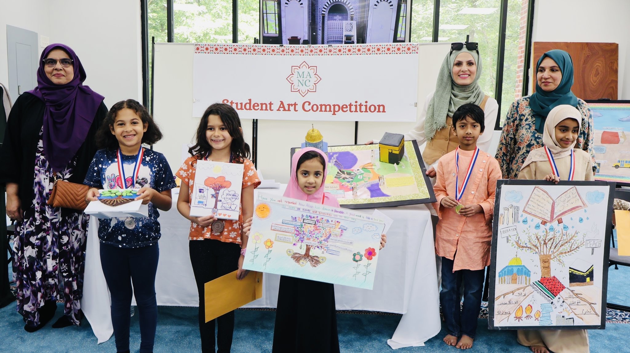Group of children and adults displaying artwork at a student art competition, with a banner in the background.