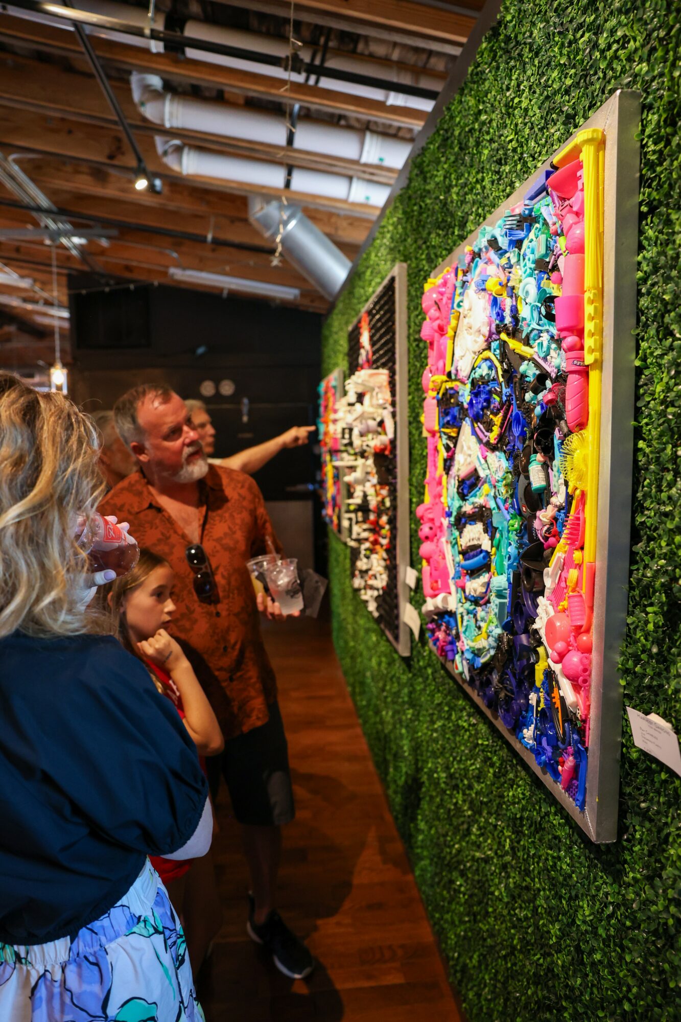 People viewing colorful abstract artwork displayed on a green wall at an art gallery, with wooden ceiling and industrial duct visible.