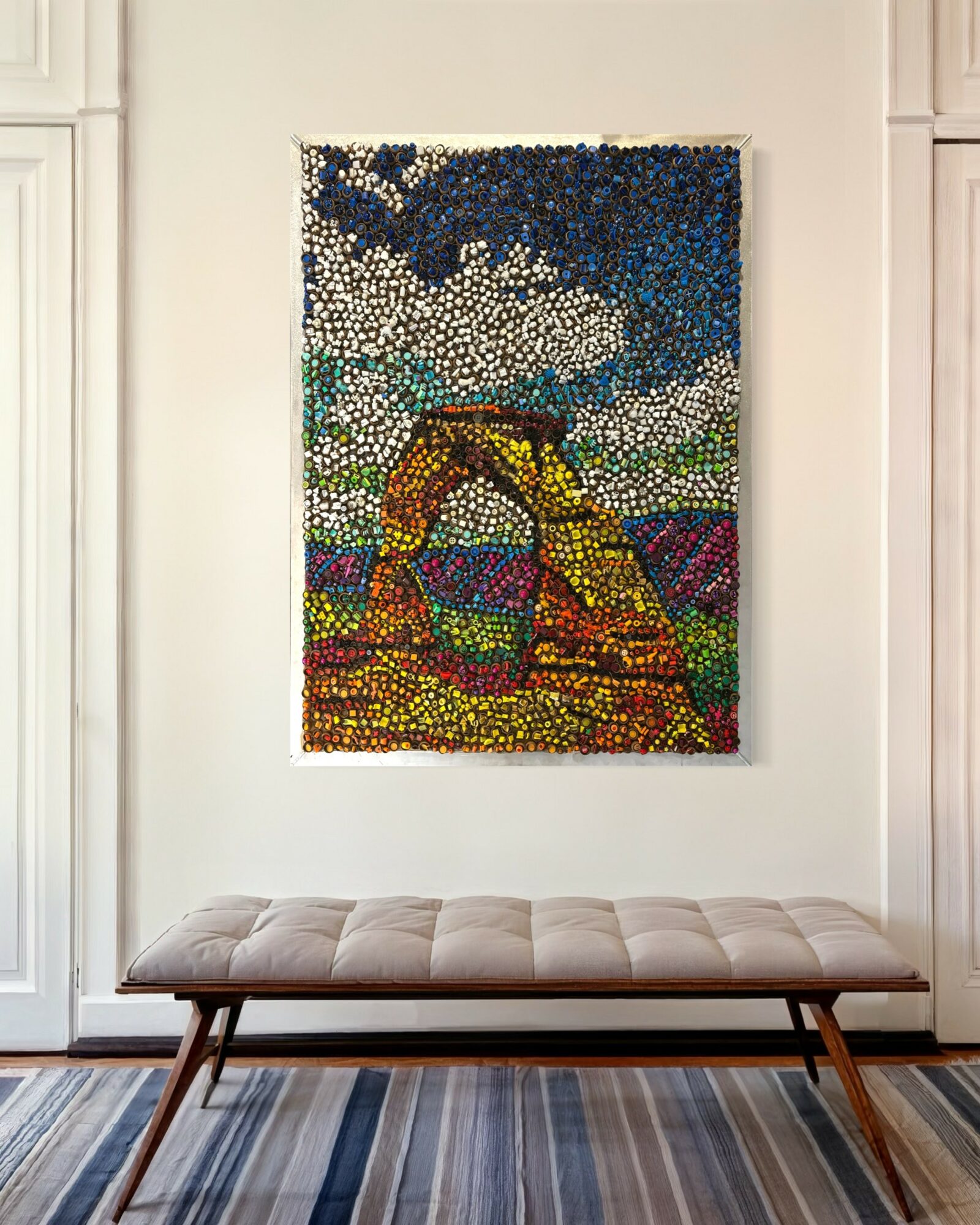 Colorful mosaic artwork depicting a natural landscape with sky, mountains, and a bridge, hanging on a white wall above a bench.