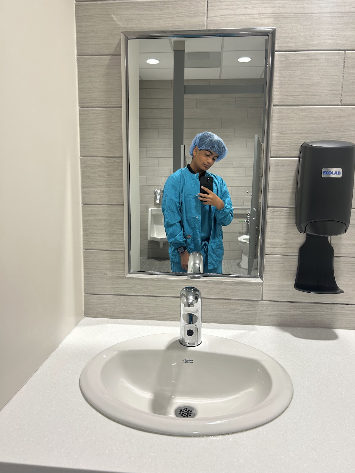 Person in blue protective clothing taking a selfie in a bathroom mirror, with sink and hand sanitizer dispenser visible.