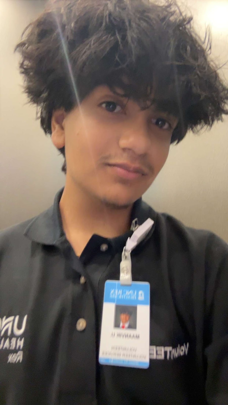 Young person with curly hair wearing a black polo shirt and an ID badge, smiling slightly.