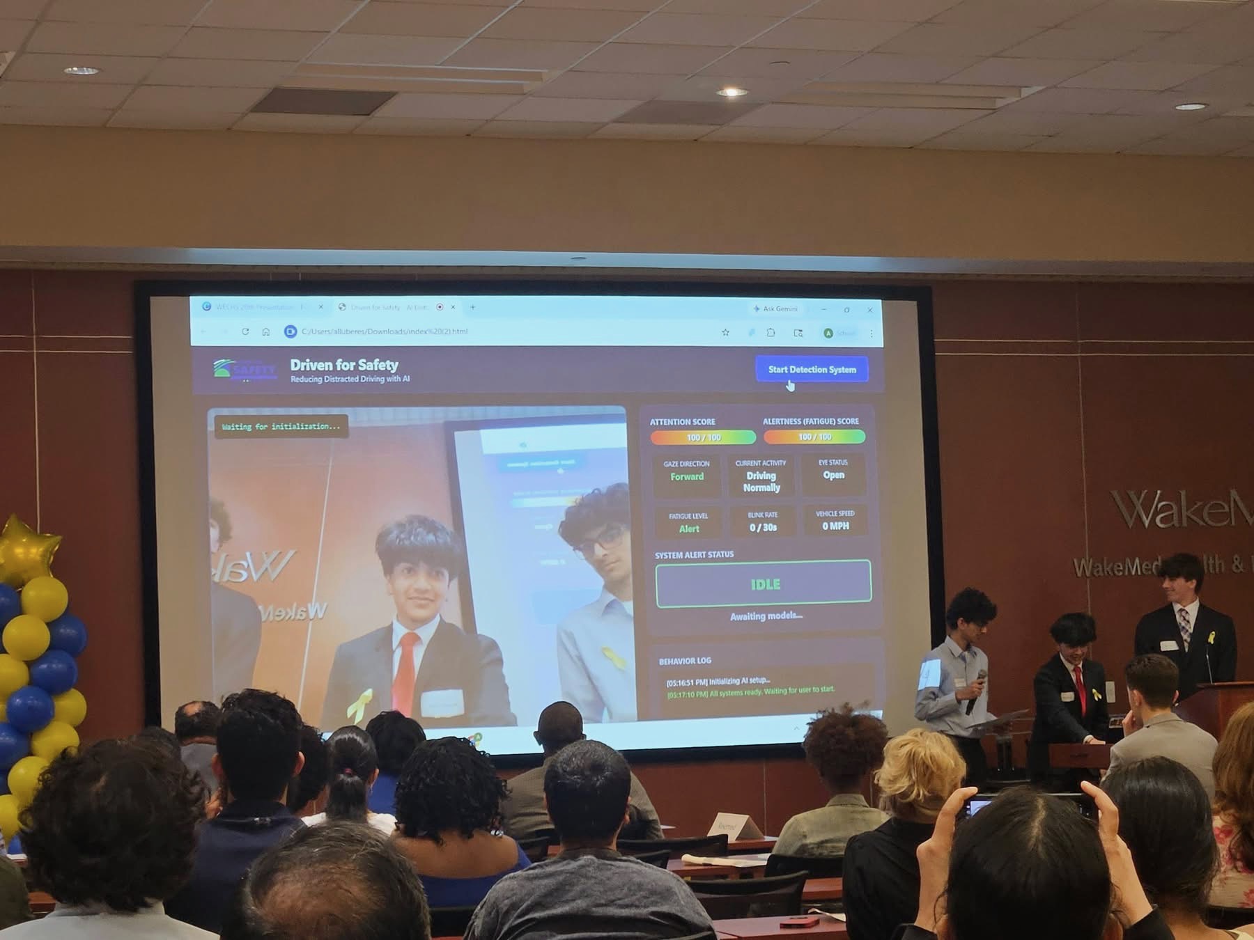 People seated in a room watching a presentation projected on a screen, with children standing to the right of the screen.