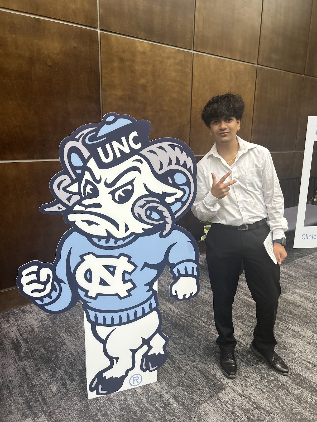 Young person in white shirt and black pants standing next to a UNC mascot cutout, making a hand gesture, in an indoor setting.