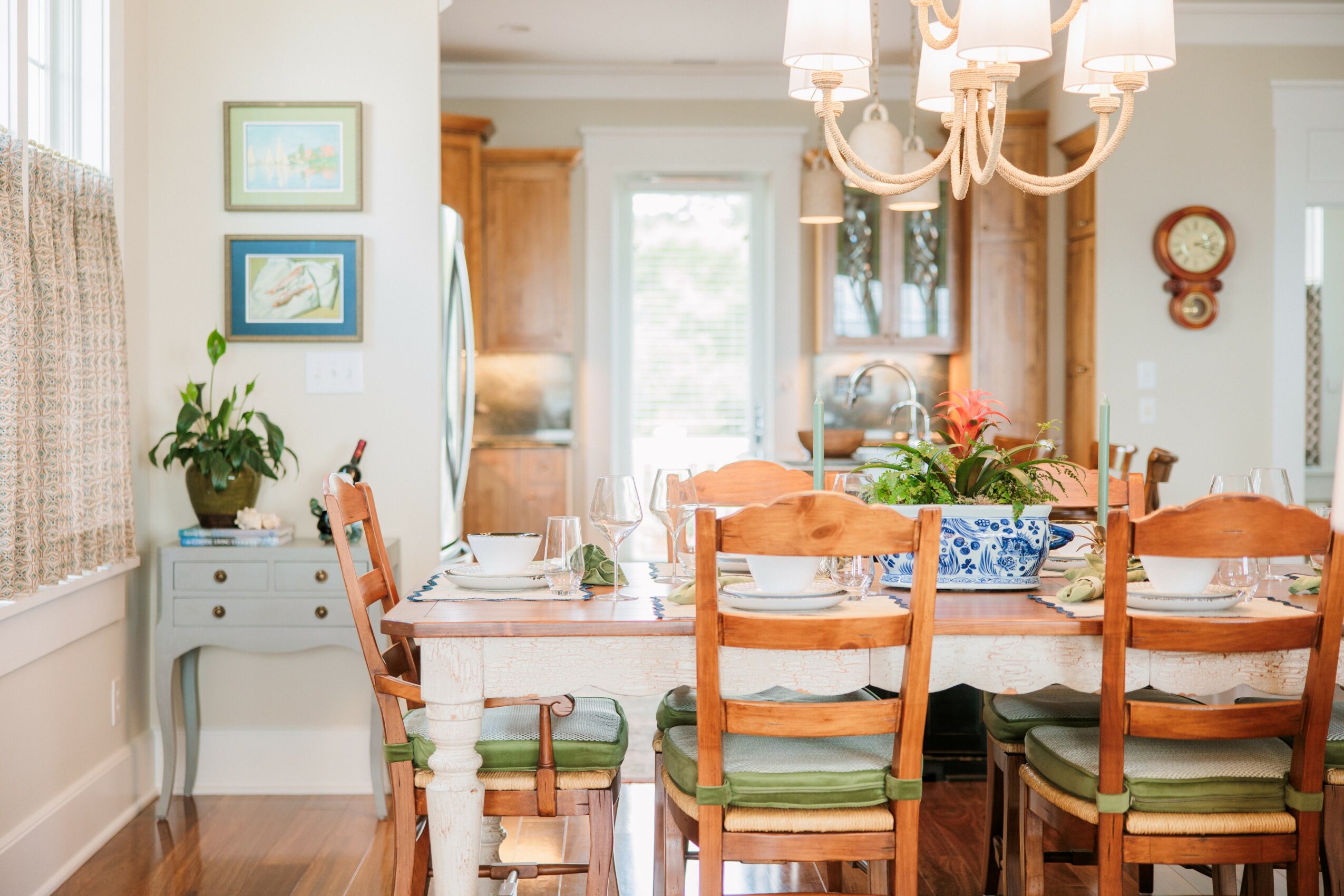 Dining room with a table, chairs, chandelier, and window with curtains, decorated with plants and artwork.
