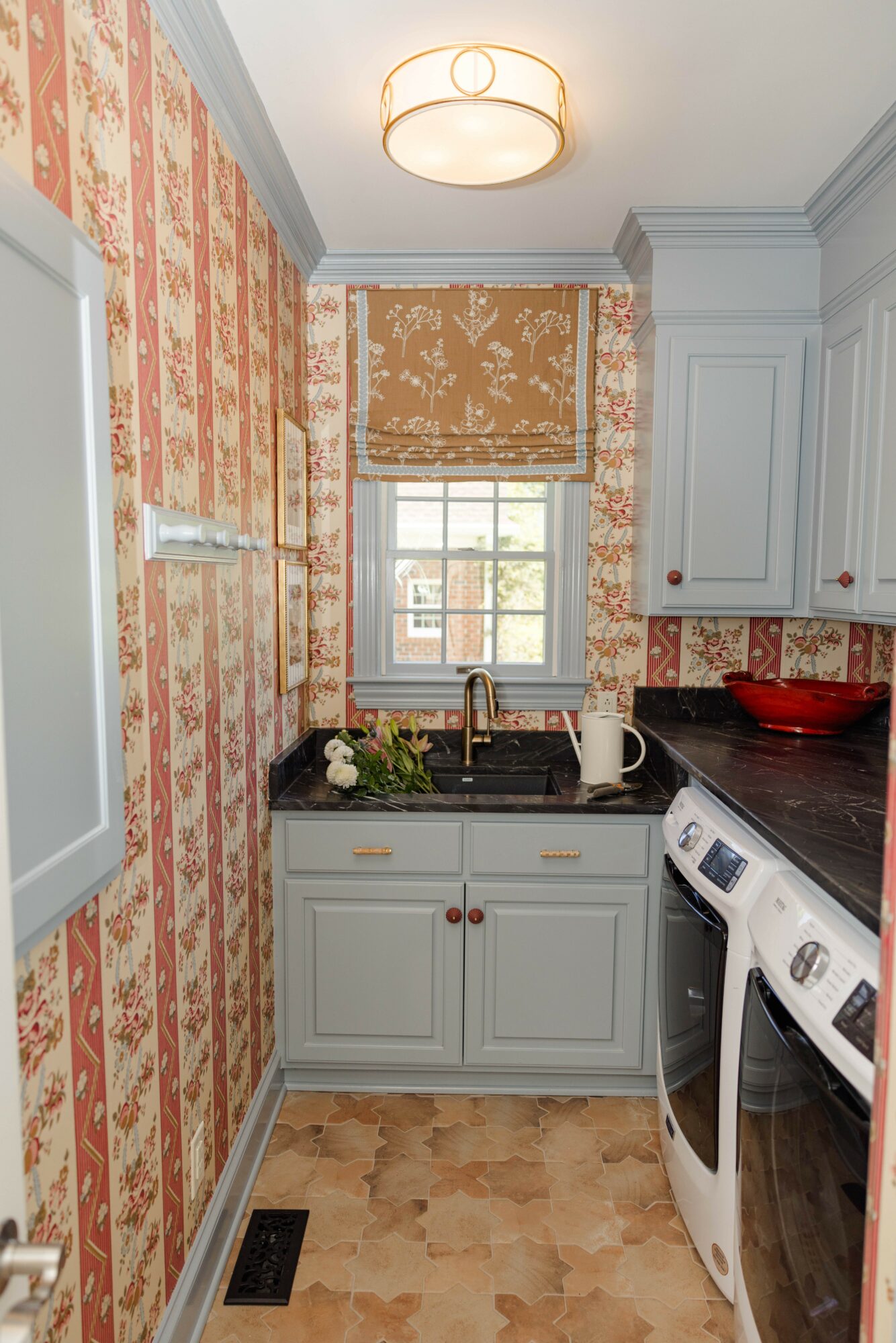 Small kitchen with patterned wallpaper, window with Roman shade, white cabinets, black countertop, and washing machine.