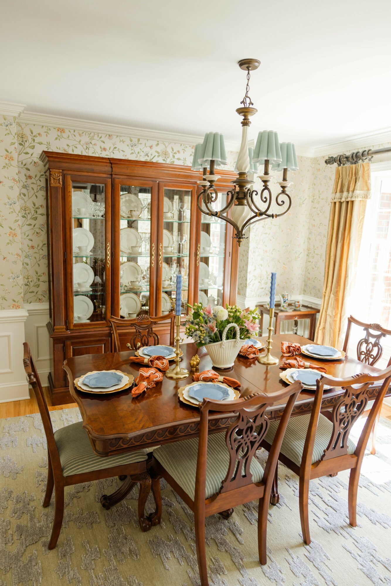 Dining room with wooden table, chairs, china cabinet, chandelier, and window with curtains, decorated with floral wallpaper.