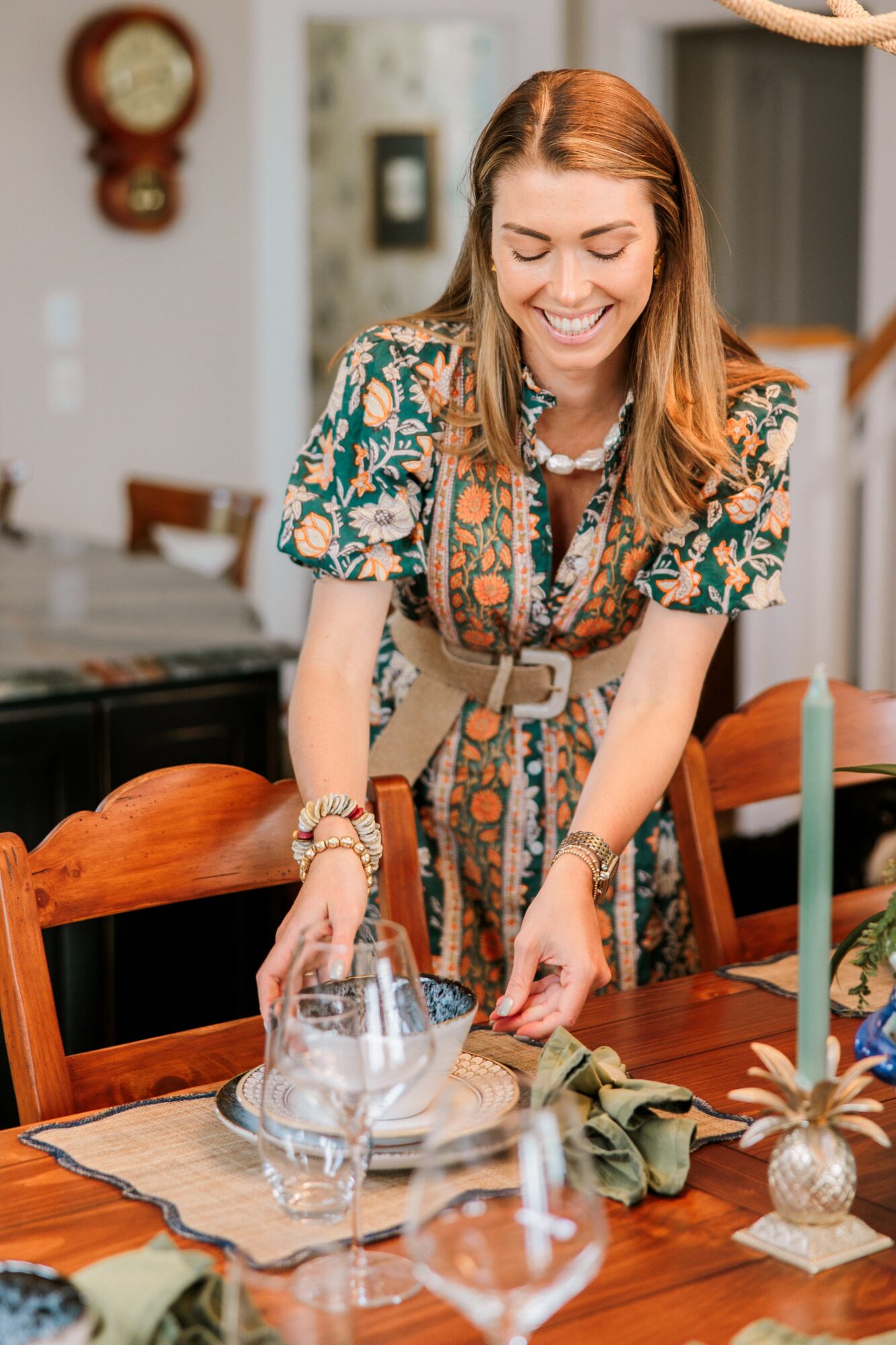 Woman with red hair smiling, setting a table with glassware and a cloth, in a decorated room.