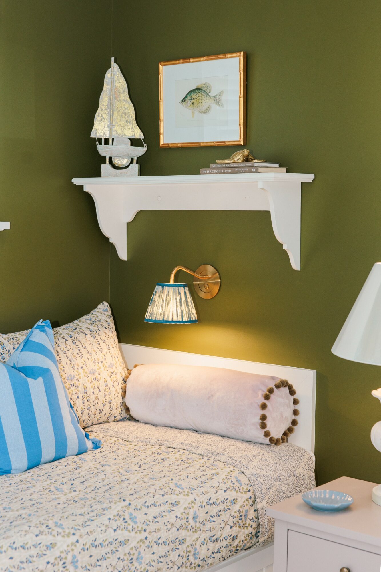 Bedroom corner with green wall, white shelf, framed fish picture, and bed with pillows and rolled blanket.