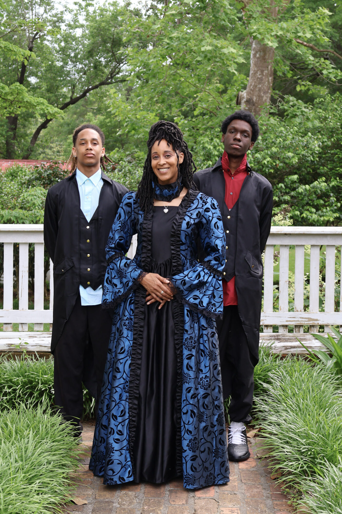Three people standing outdoors on a pathway with green trees and grass, dressed in formal attire, smiling at the camera.