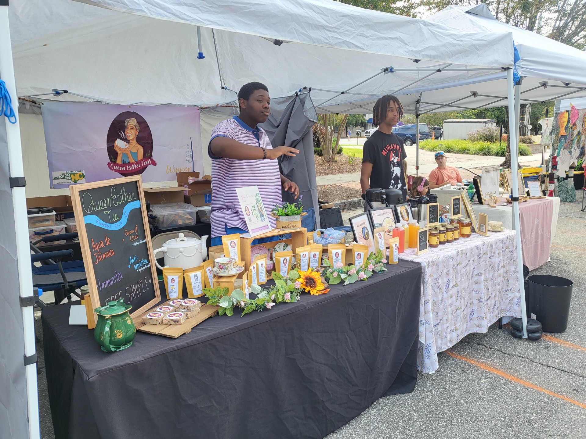 Two people stand behind a table at an outdoor market stall with various jars, signs, and decorations, under a white canopy.