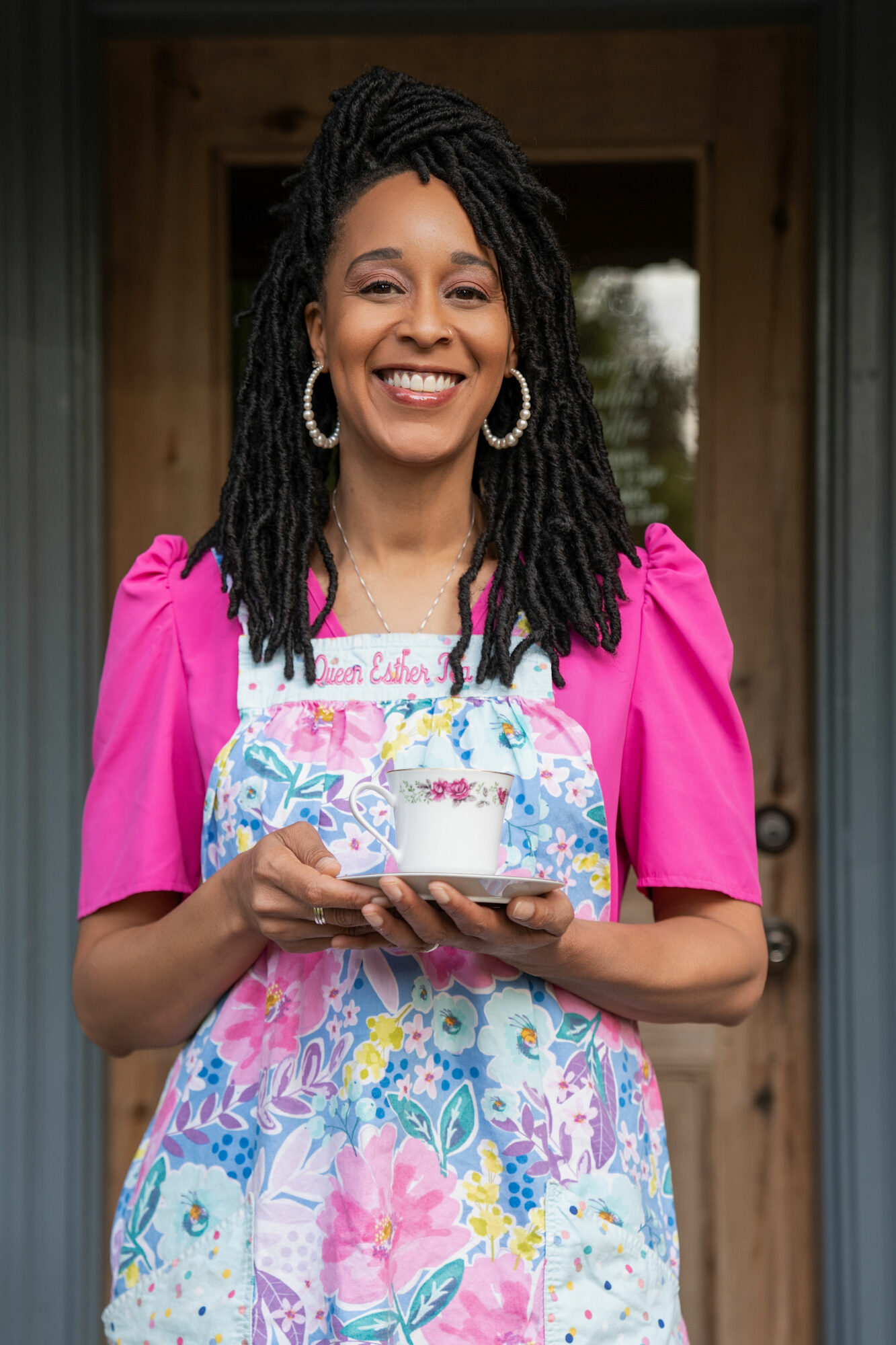 Smiling woman with dreadlocks holding a cake, standing in front of a wooden door, wearing a pink top and floral apron.