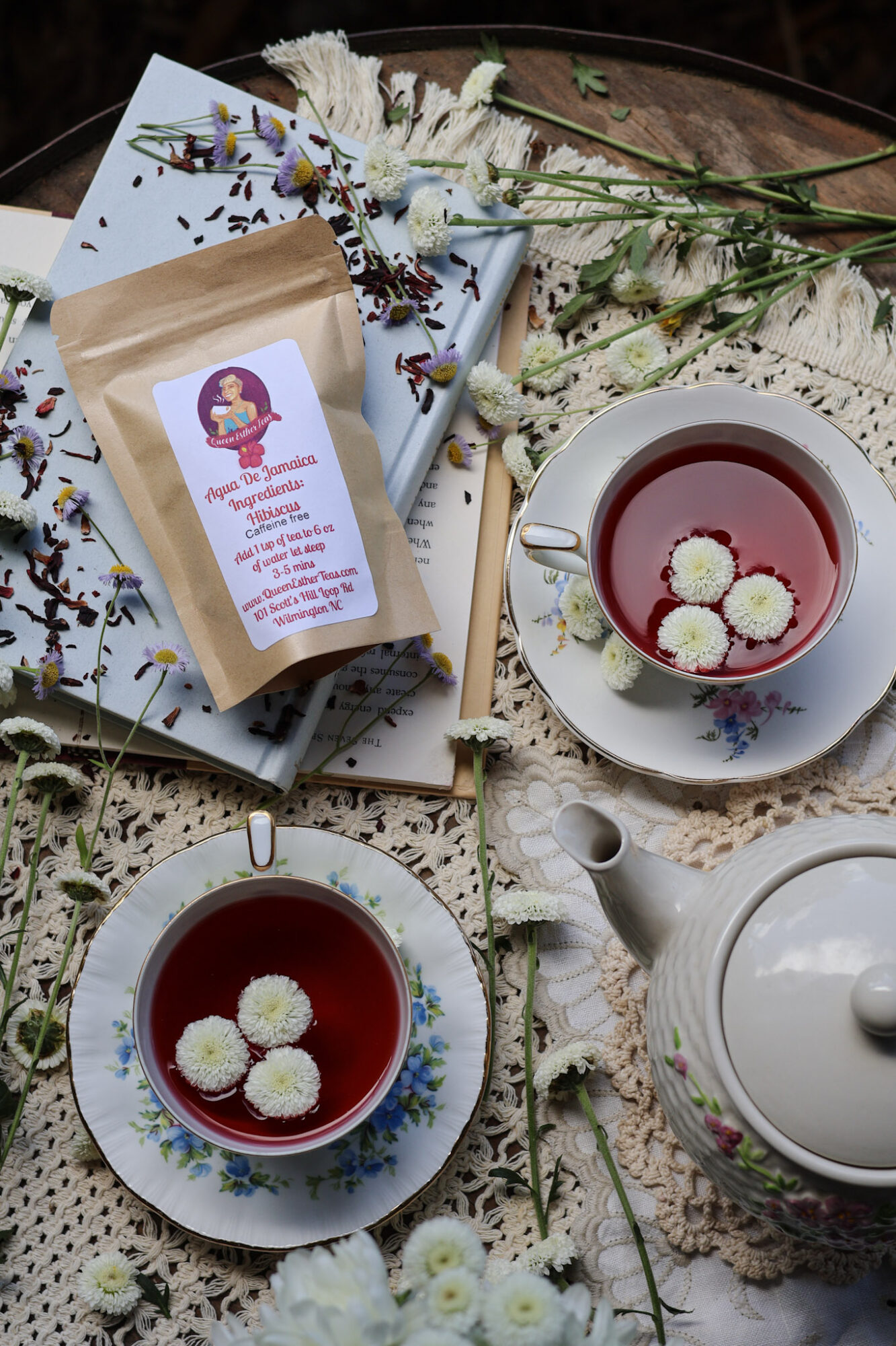 Tea setup with two cups of tea, a teapot, flowers, and a package on a lace cloth, with books and flowers around.