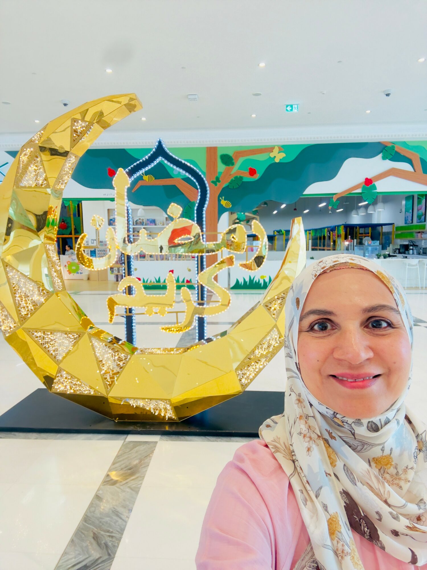 Woman taking selfie in front of a decorative crescent moon and Arabic script display, with colorful background and indoor setting.