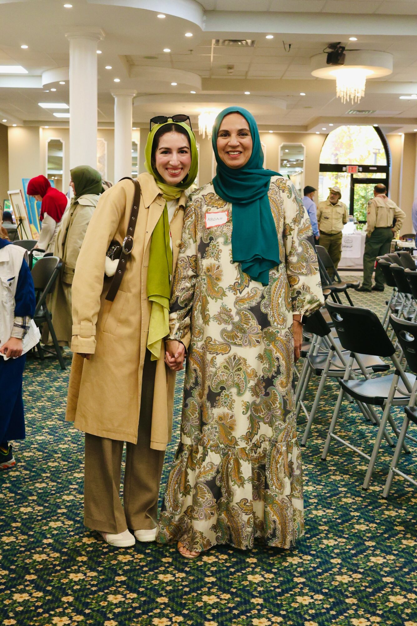Two women standing together in a large indoor space, smiling, with chairs and people in the background.
