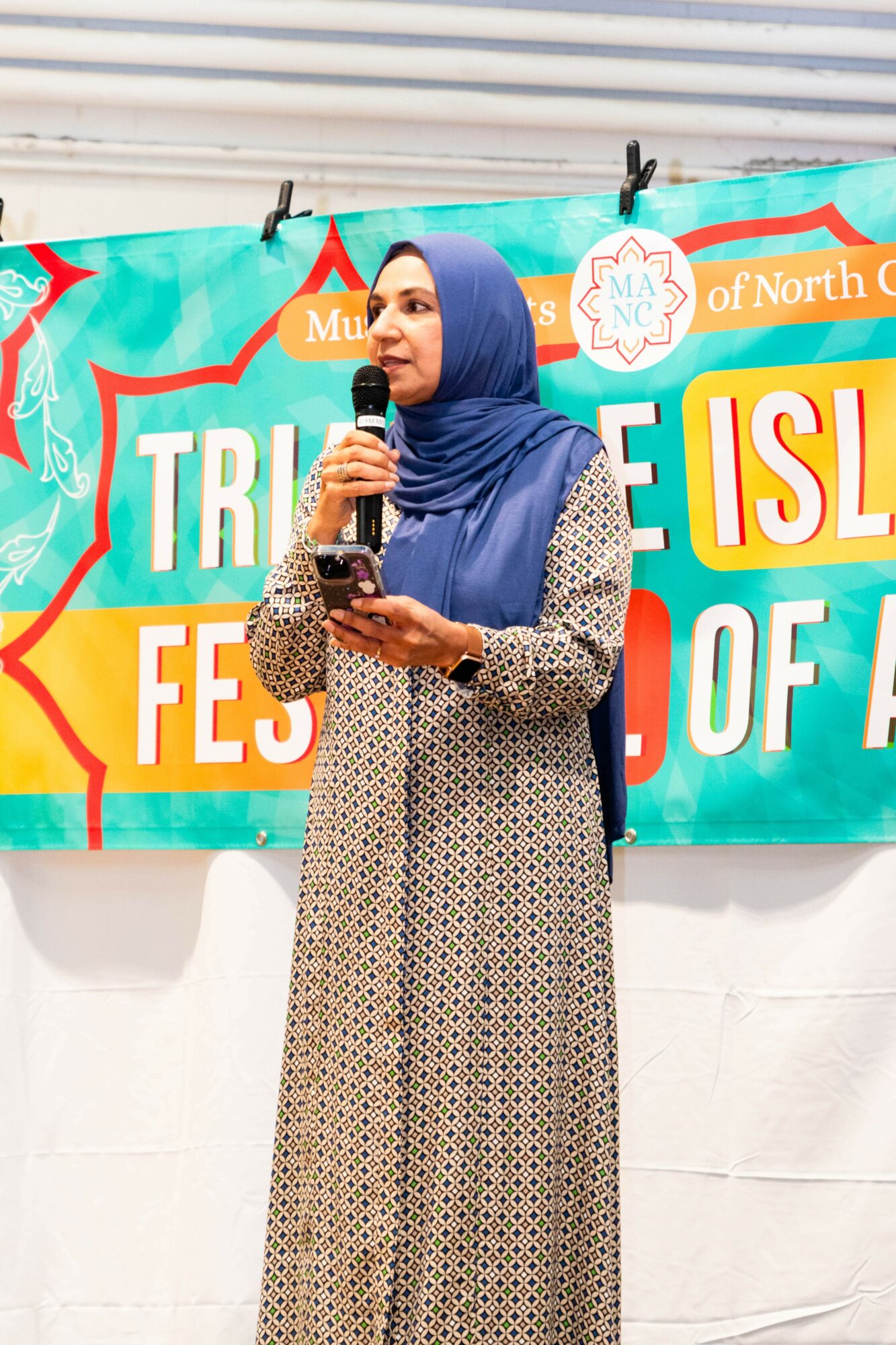 Woman in blue hijab and patterned dress speaking into a microphone at an outdoor event, with a colorful banner behind her.