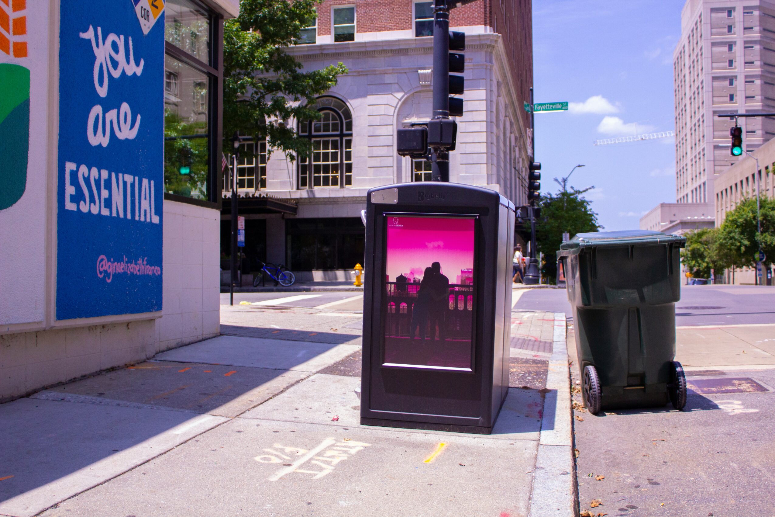 City street corner with a digital kiosk, trash bin, and a large blue sign on the wall, with buildings and trees in the background.