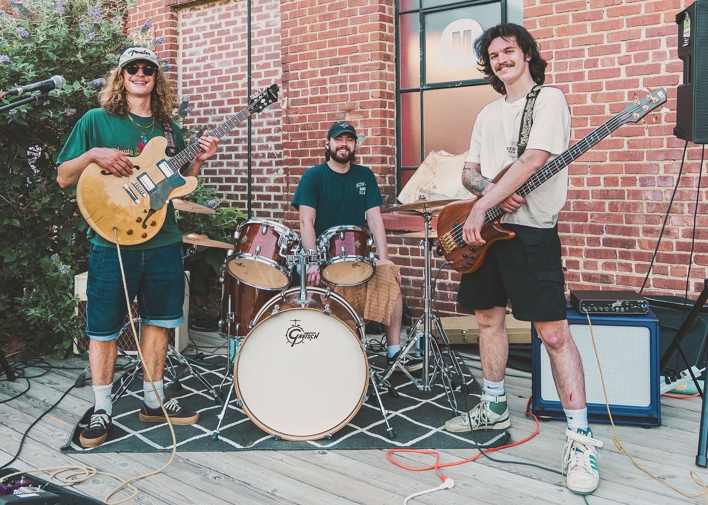 Three musicians with guitars and drums perform outdoors on a brick patio, smiling and playing instruments.