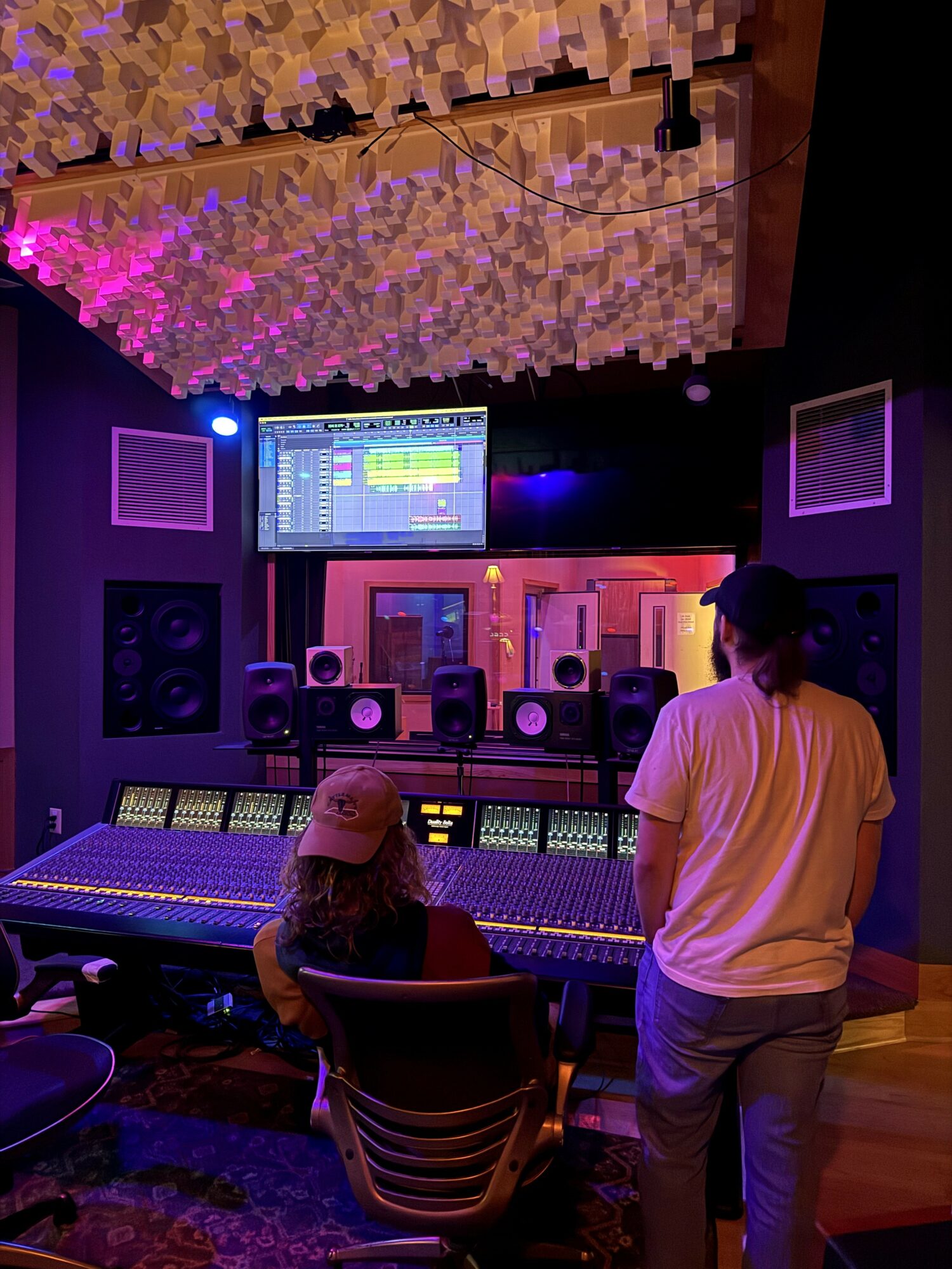 Two people in a recording studio with a large mixing console, speakers, and a screen, one sitting and one standing, ceiling with acoustic panels.