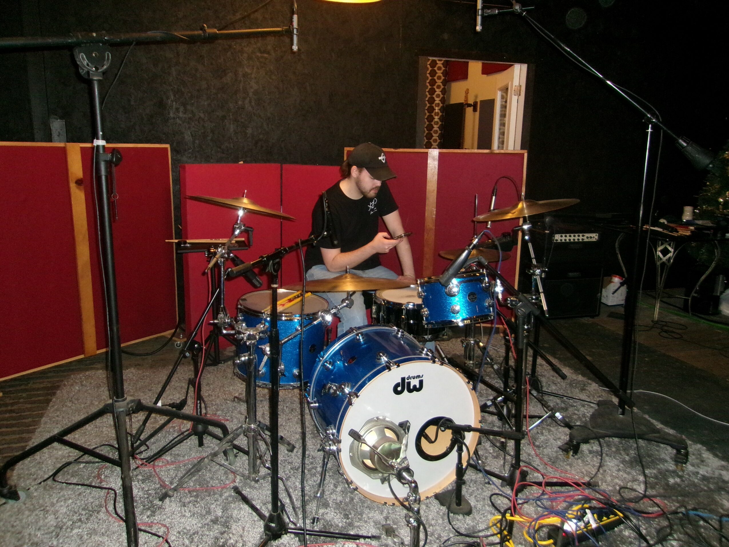 Person sitting behind a blue drum set on a stage with red and black background, surrounded by microphones and equipment.