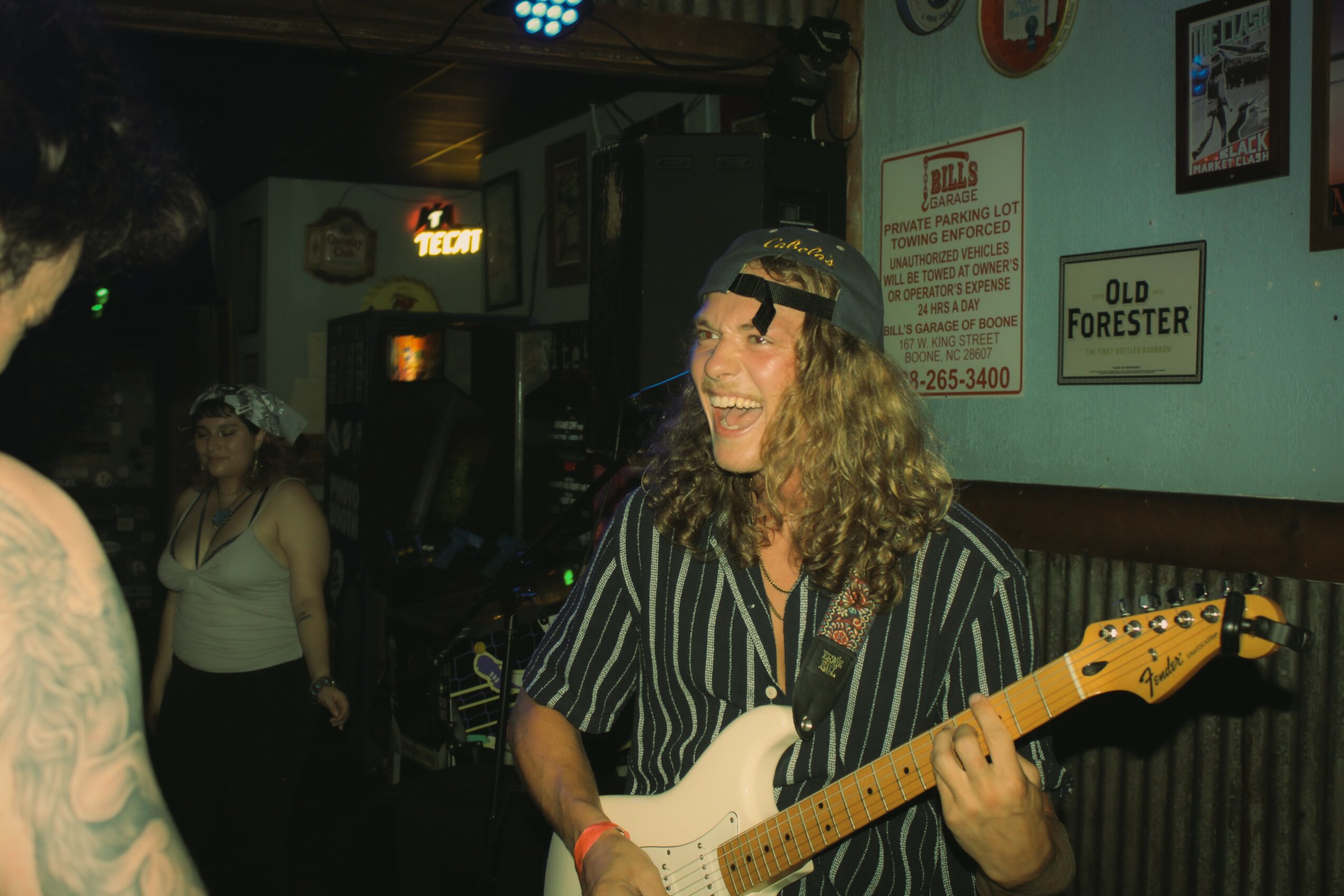 Person with curly hair playing an electric guitar, smiling, wearing a striped shirt and a headband, in a bar setting.