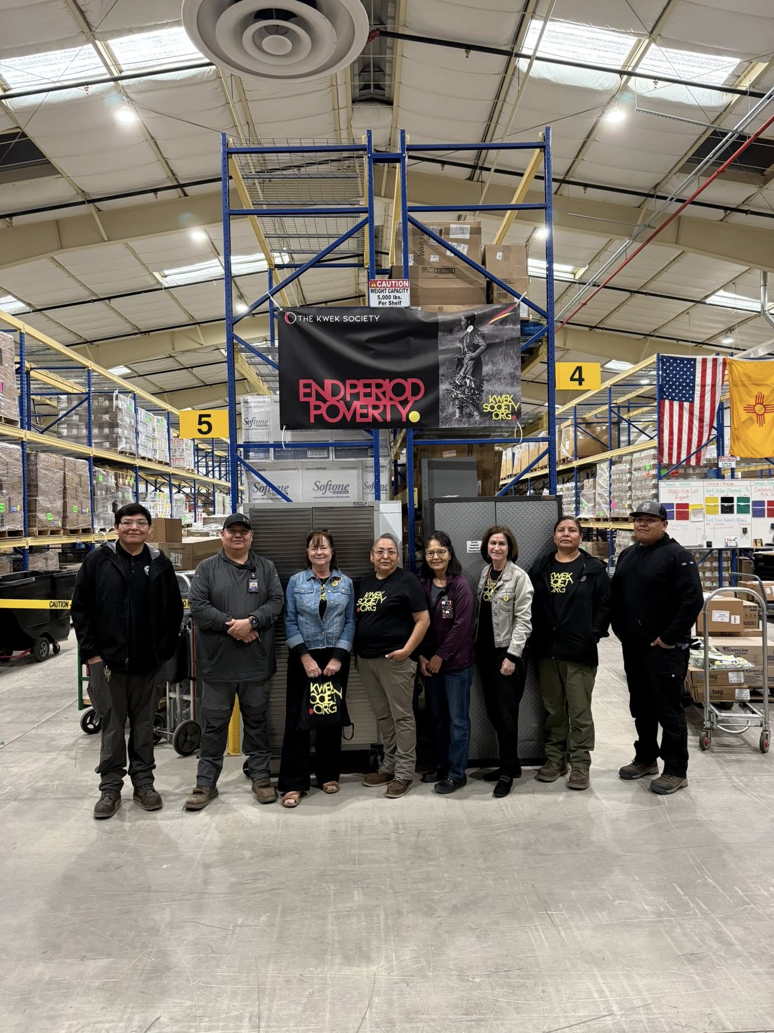 Group of seven people standing in warehouse with scaffolding and American flags in background.
