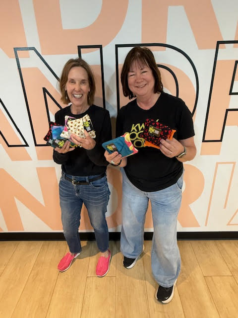 Two women standing indoors, smiling, holding colorful socks, with large text background, wooden floor, casual clothing.