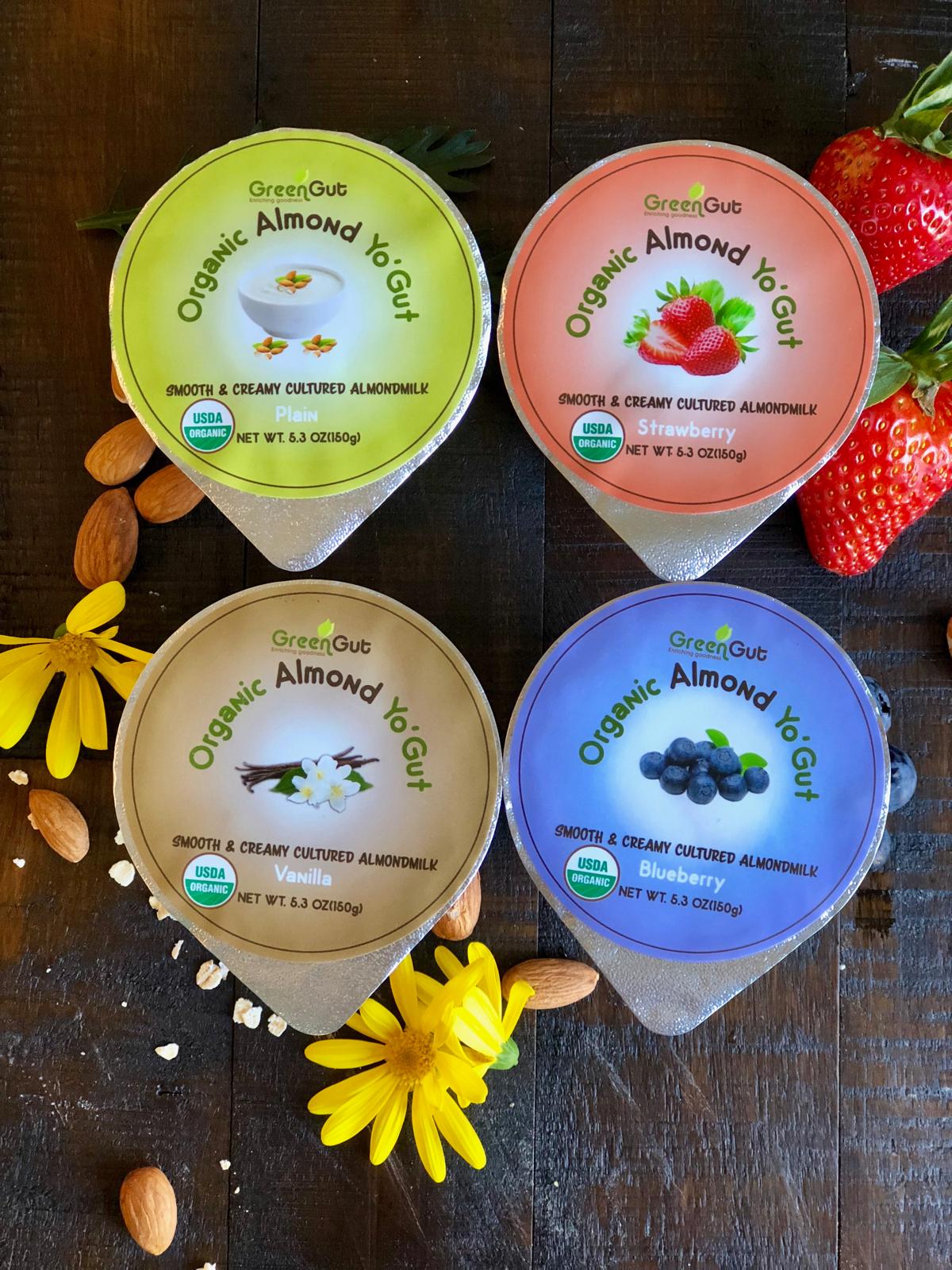 Four containers of organic almond yogurt with different flavors, surrounded by strawberries, almonds, and yellow flowers on a dark surface.