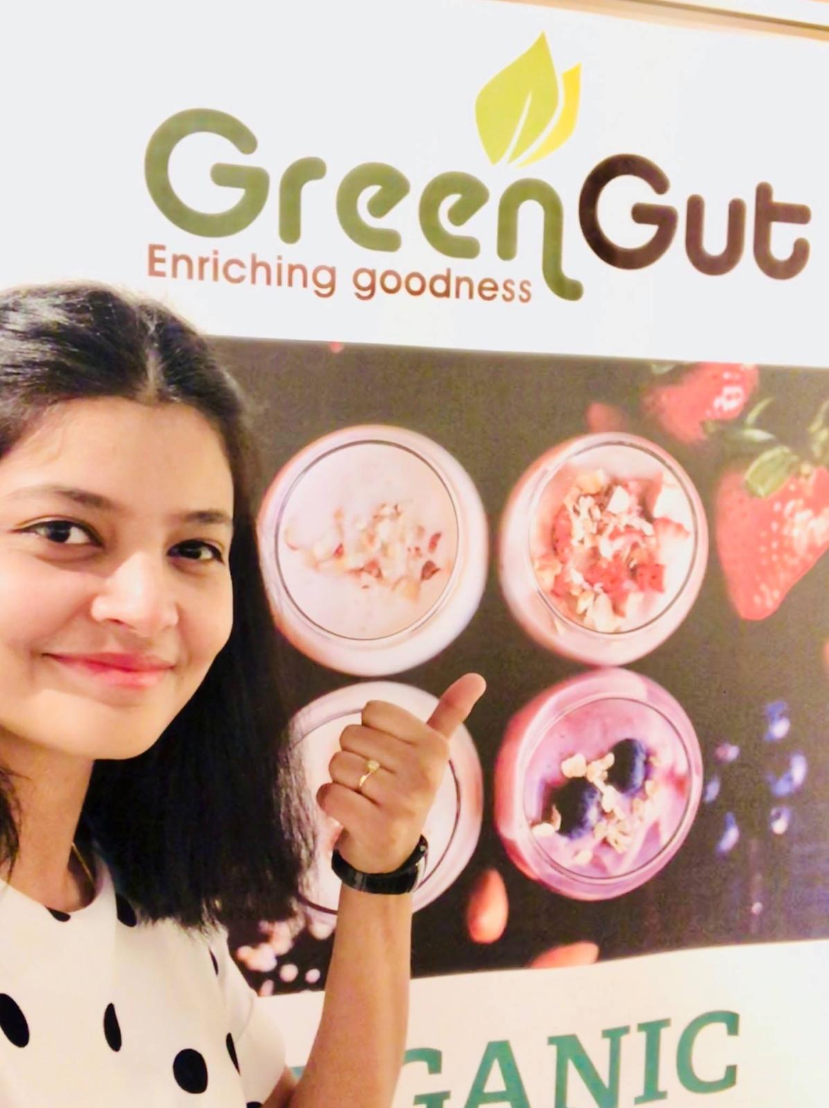 Young girl with dark hair smiling and giving a thumbs-up in front of a sign for Green Gut, an organic product brand.