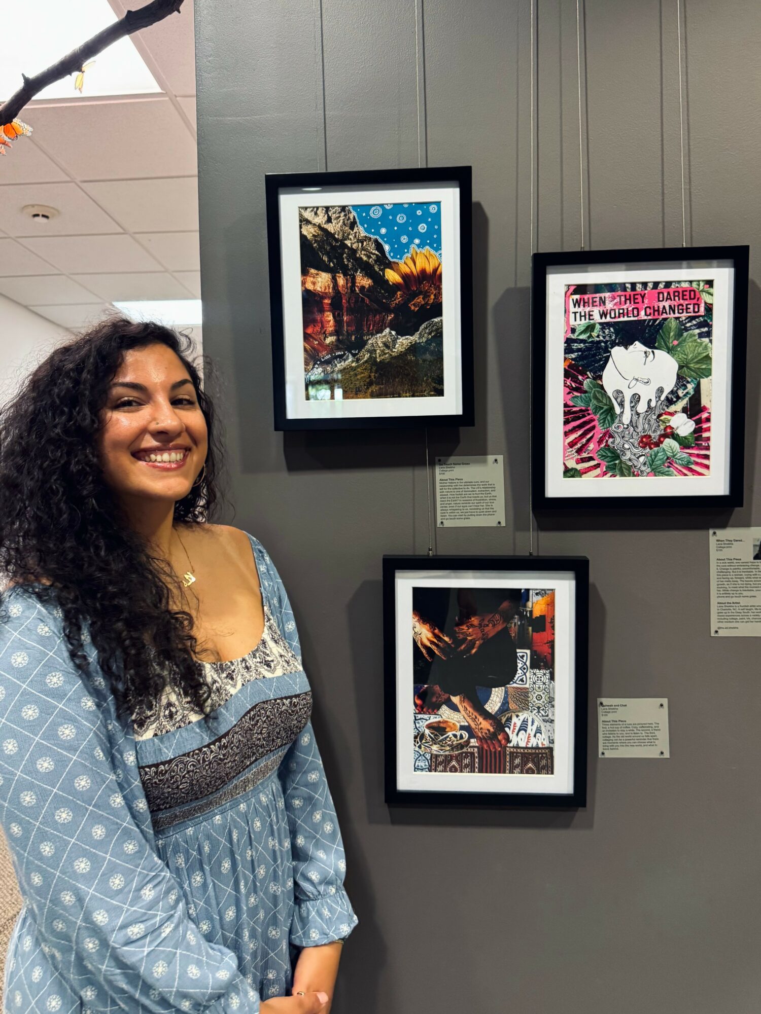 Smiling woman with long curly hair standing in front of framed artwork on a gray wall.