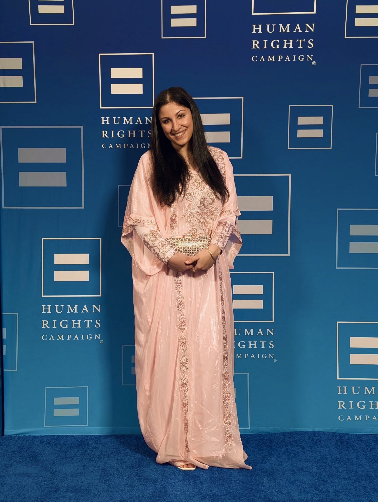 Woman in pink dress holding a clutch, standing in front of a blue backdrop with Human Rights Campaign logos.