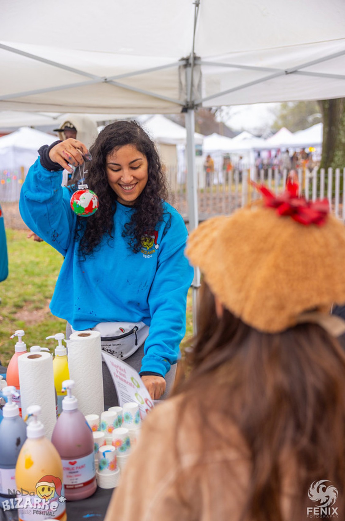 Young woman in blue sweatshirt smiling at a table with colorful bottles, outdoors under a white canopy, with a person wearing a straw hat nearby.