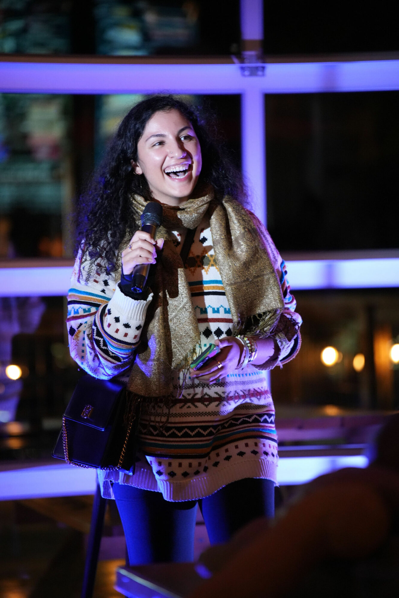 Young woman smiling and holding a microphone indoors at night, wearing a patterned sweater and scarf.